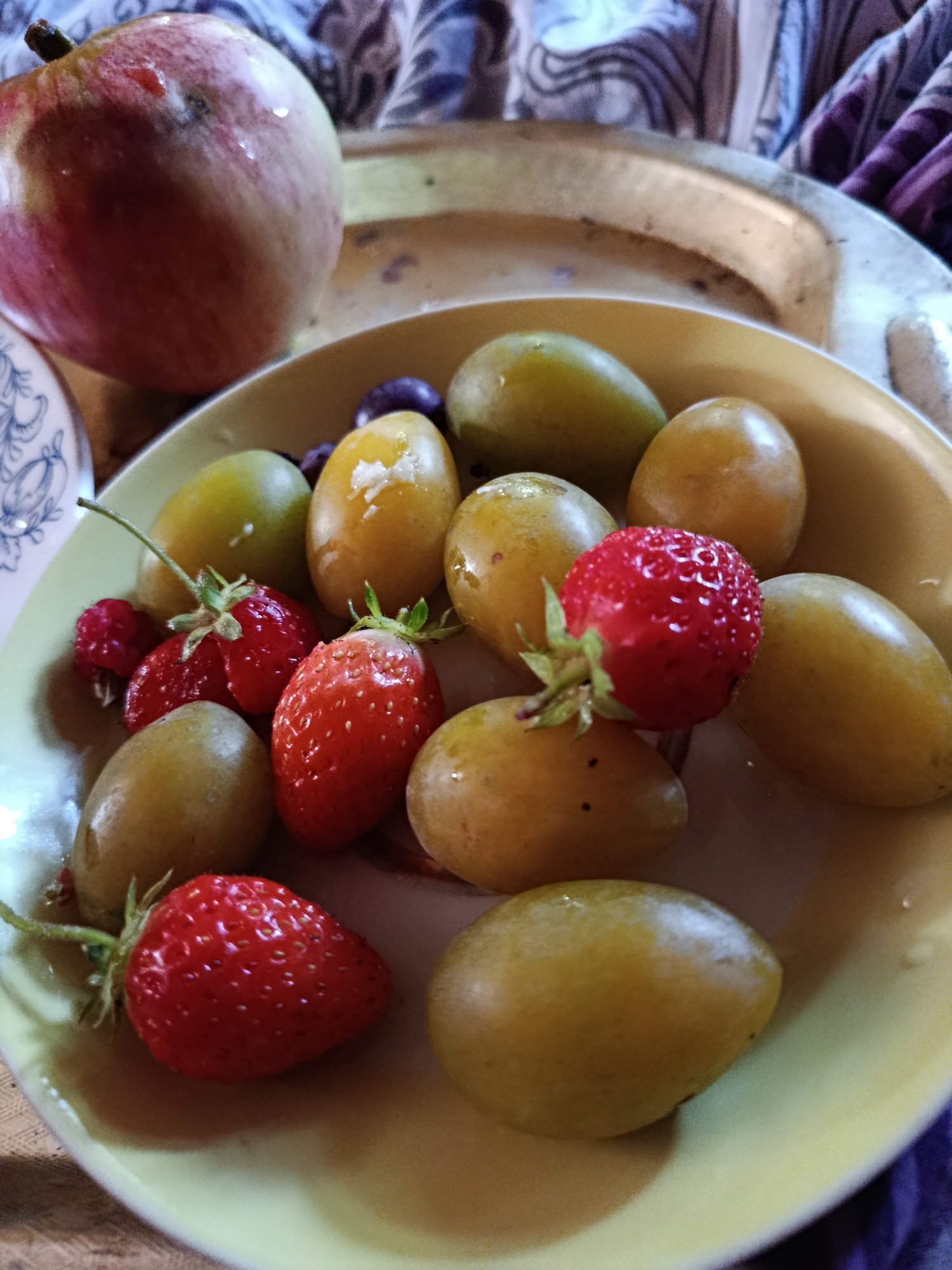 An overhead shot shows a plate of fruit, including an apple, strawberries, and small yellow plums. The plate is shallow and yellow. A few dark purple plums are visible in the background of the plate. The apple is on the left side of the image and is red and yellow. The strawberries are red and have their green tops. The yellow plums are oval-shaped. The fruit appears to be freshly picked and is glistening with water droplets. The image is taken in the city of Верея, Russia. The background is blurred, but it appears to be a tablecloth with a patterned design. The lighting is natural, and the overall impression is one of freshness and abundance.