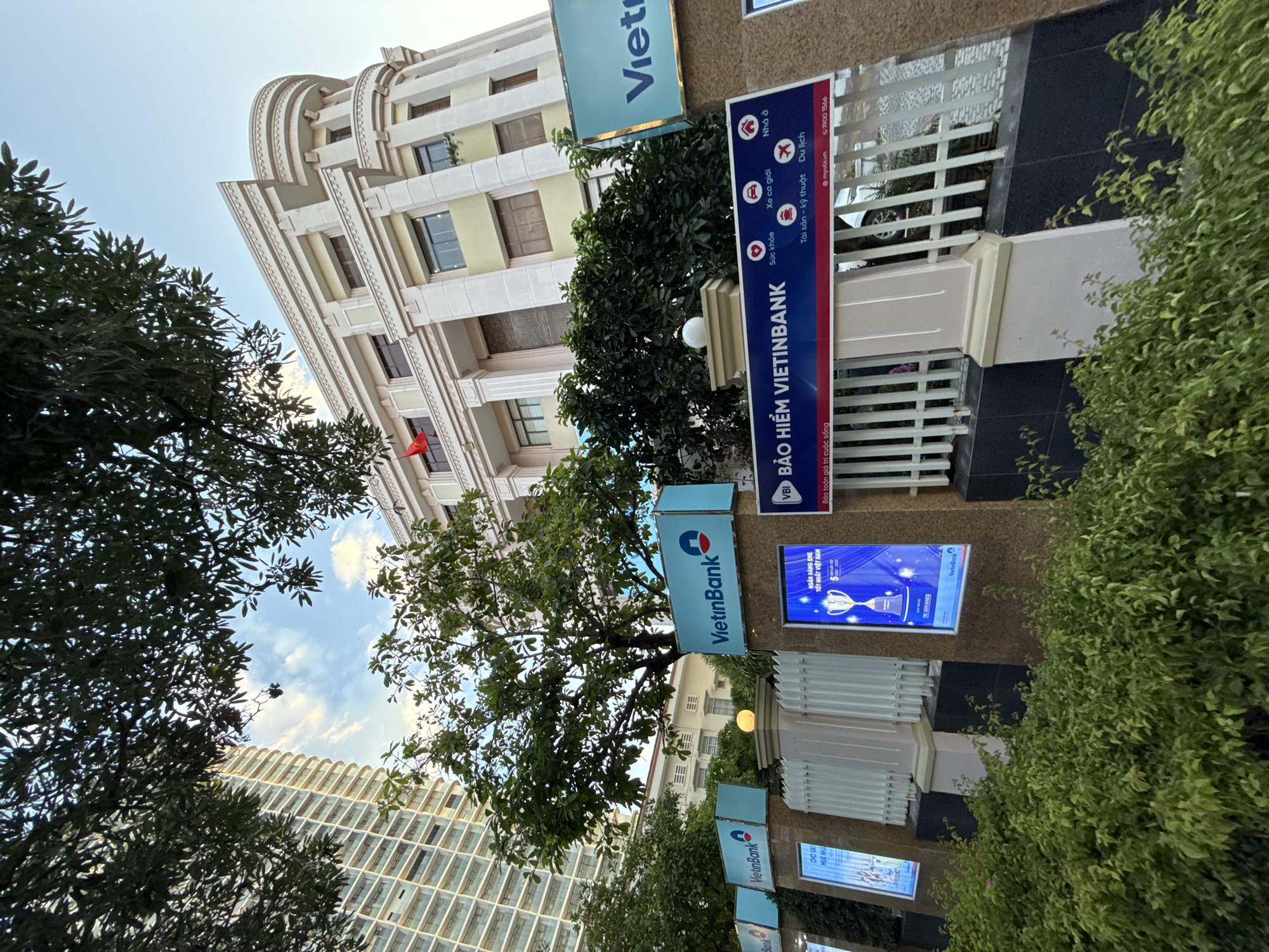 The image shows a modern building with a curved facade, identified by signage as belonging to VietinBank, a Vietnamese bank. The building appears to be located in Nha Trang, Vietnam, as indicated by the Vietnamese text on the signage. In the foreground, there is a fence with several illuminated advertisements displaying bank services and awards. Lush green bushes line the bottom of the image. Trees with dense foliage are visible in front of and around the building. A tall, modern high-rise building is partially visible to the left of the main VietinBank building. The sky is partly cloudy, suggesting it is daytime.