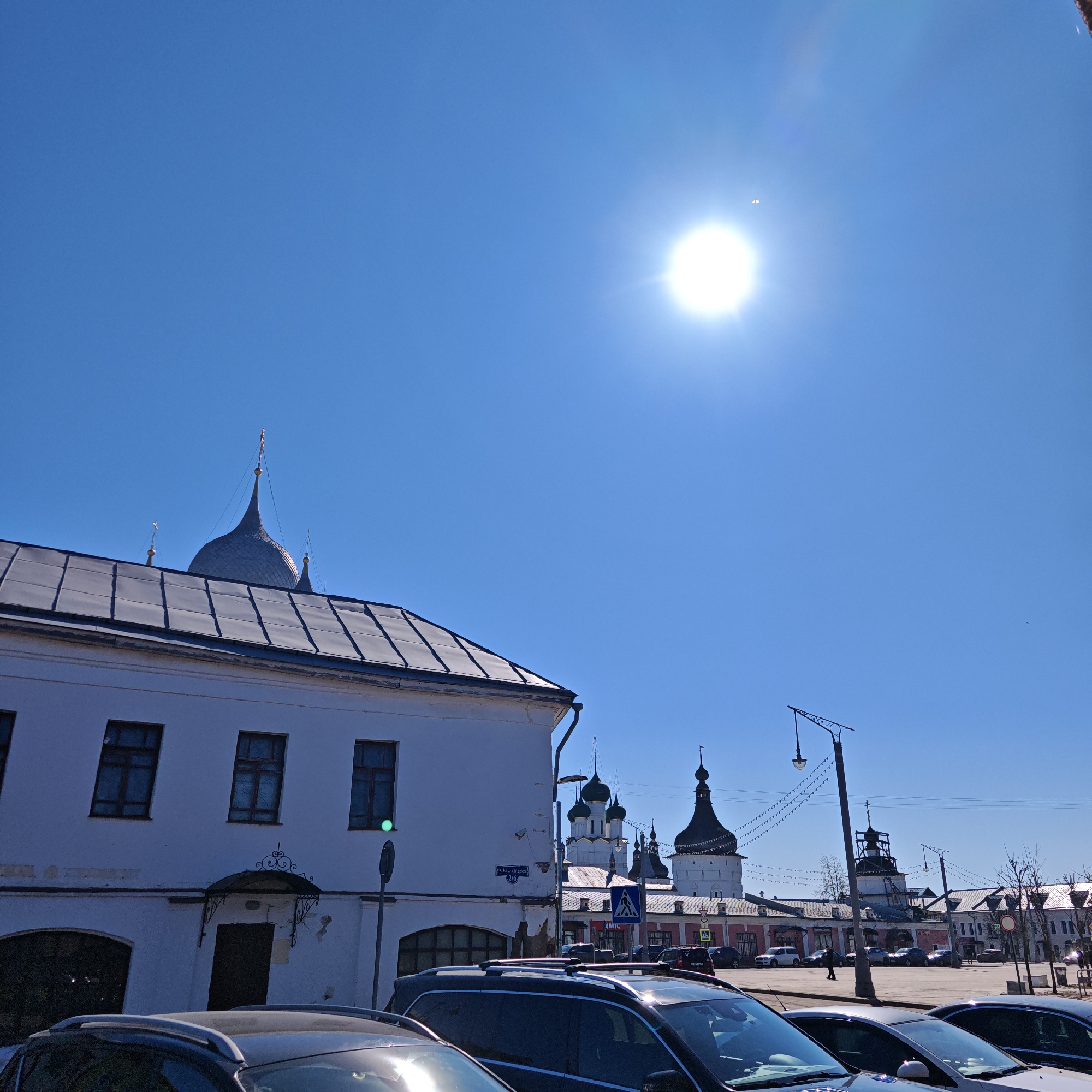 The photograph depicts a street scene in Rostov, Russia, under a clear, bright sky with a strong sun. In the foreground, several parked cars are visible. Behind the cars, a white, two-story building with a grey roof is situated to the left of the frame. In the background, there are various towers and domes characteristic of Russian Orthodox architecture. These structures suggest the presence of a monastery or cathedral complex. A pedestrian crossing sign is visible. A lamp post is present, and overhead festoon lighting stretches across the street. The overall impression is of a sunny day in a historic urban environment.