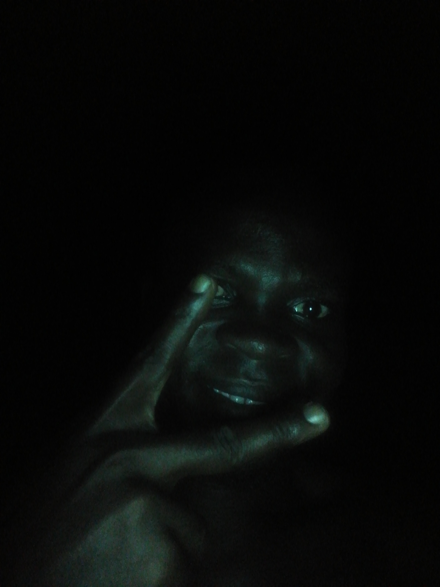 A close-up, low-light photograph shows a dark-skinned person, possibly male, with a smile. The person's left hand is raised, with the index and middle fingers forming a "peace" or "victory" sign. The background is predominantly black. There is no information provided that indicates the photo's location is Ikorodu, Nigeria.