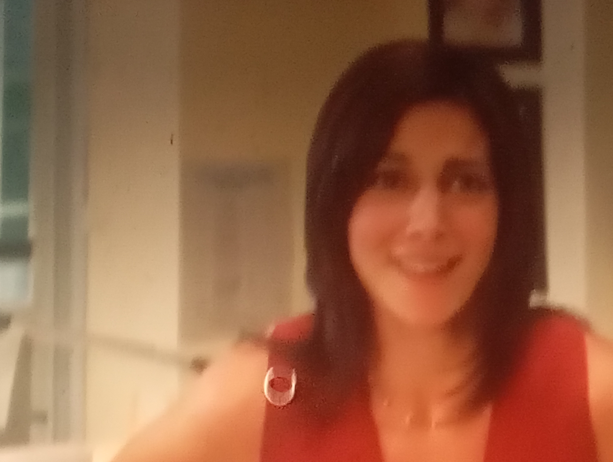 This is a photograph of a woman with dark hair, wearing a red top. She is smiling and looking towards the camera. The background is blurred but appears to be indoors, with warm-toned walls. There are some indistinct objects in the background, possibly framed pictures on the wall. The lighting suggests it might be daytime or well-lit indoors. The image quality is low, with a grainy appearance. The woman is wearing a small, circular brooch on her red top. The setting appears to be a domestic environment. No text is visible in the image.