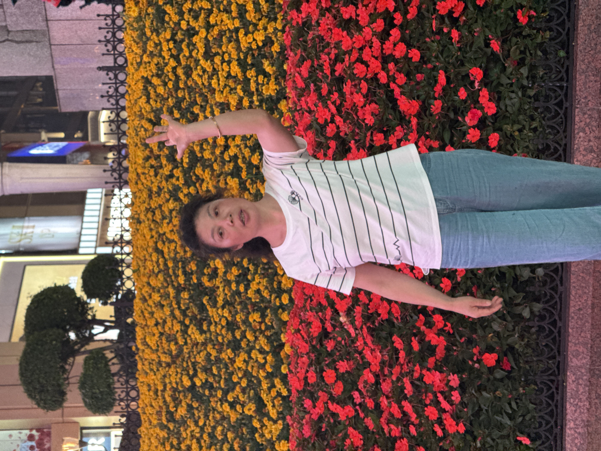 A woman stands in front of a dense display of flowers in Yuzhong District, China. To her left are yellow flowers and to her right are red flowers. She is wearing a striped white t-shirt and light blue jeans. Her right arm is outstretched towards the yellow flowers with her fingers splayed. Her left arm is outstretched toward the red flowers, and her fingers are straight. In the background, a building is visible, along with manicured trees and a black wrought iron fence.
