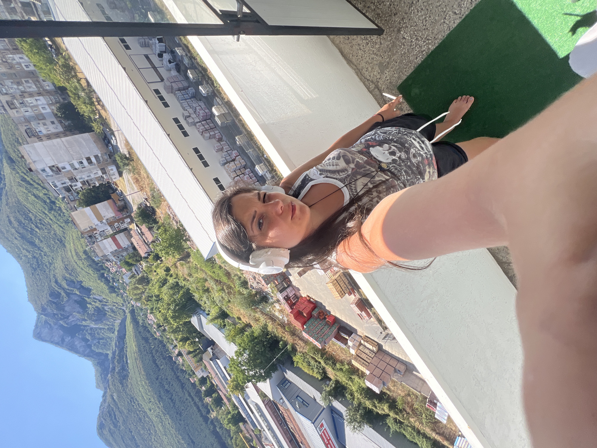 A woman is taking a selfie from a balcony in Vratsa, Bulgaria, overlooking a cityscape with mountains in the background. She is wearing white headphones, a black tank top with a skull print, and black shorts. She is barefoot and holding a cigarette. The balcony has a white railing and a green mat. In the background, there are buildings, factories, and stacked materials, indicating an industrial or commercial area. The sky is clear and blue, and the sun is shining brightly.
