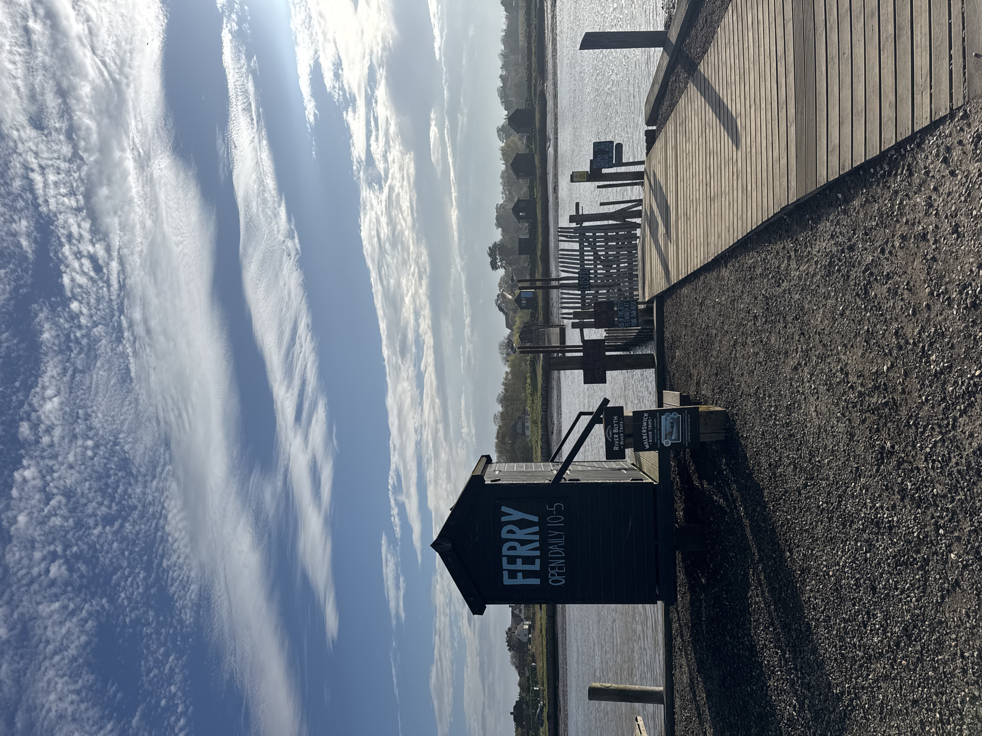 The scene captures the Walberswick Ferry station in East Suffolk, United Kingdom. A small wooden building stands prominently with "FERRY" painted on its side, accompanied by the text "OPEN DAILY 10-5". Beside the building are wooden posts and a section of wooden fencing leading to a wooden pier extending into the water. Gravel covers the ground leading to the pier. The sky is bright blue with streaky, cloud formations. The ferry station borders a body of water with land visible in the distance. Signage with information about "RIVER BLYTH - RIVER TRIPS -" and "WALBERSWICK" is visible beside the building.