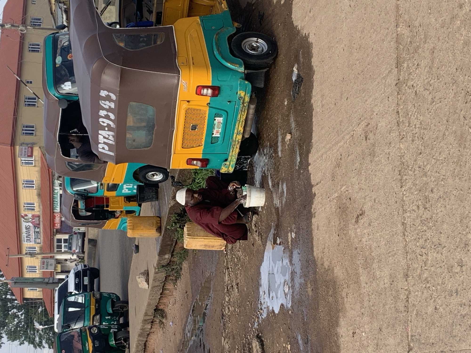 The image depicts a street scene in Gura Top, Nigeria, with several auto-rickshaws, also known as Keke Napeps, parked. In the foreground, a man wearing a maroon robe and a white cap is squatting by the side of the road, washing his hands in a white bucket. The ground is muddy and wet, with puddles of water scattered around. Two yellow plastic containers are visible nearby. Behind the man, the rear of a yellow and teal Keke Napep is prominent, displaying the license plate "TNK-488VT". Further back, more Keke Napeps are parked, some green and others a combination of brown and yellow. A white SUV is parked on the right side of the road. In the background, a two-story building with a red roof stands, featuring signs for "DUNAMIS" and "LA MED PHARMACY LTD." The environment appears to be an urban setting, likely a commercial or transportation hub, with evidence of recent rain or water spillage.