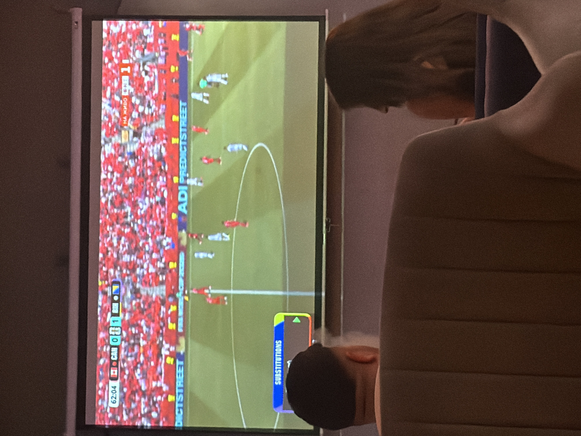 The image shows a projection screen displaying a football match, likely in a sports bar or public viewing area in Plovdiv, Bulgaria. The score on the screen indicates Canada (CAN) with 0 goals and Bosnia and Herzegovina (BIH) with 1 goal at the 62:04 minute mark. The background of the projection is filled with a crowd wearing red, suggesting it's a significant match for the Canadian team. In the foreground, the backs of two people are visible, a man on the left and a woman on the right, seated on furniture, watching the screen. The environment appears to be dimly lit, consistent with a viewing area for a projected screen. A graphic element labeled "SUBSTITUTIONS" is visible in the lower left corner of the projection.