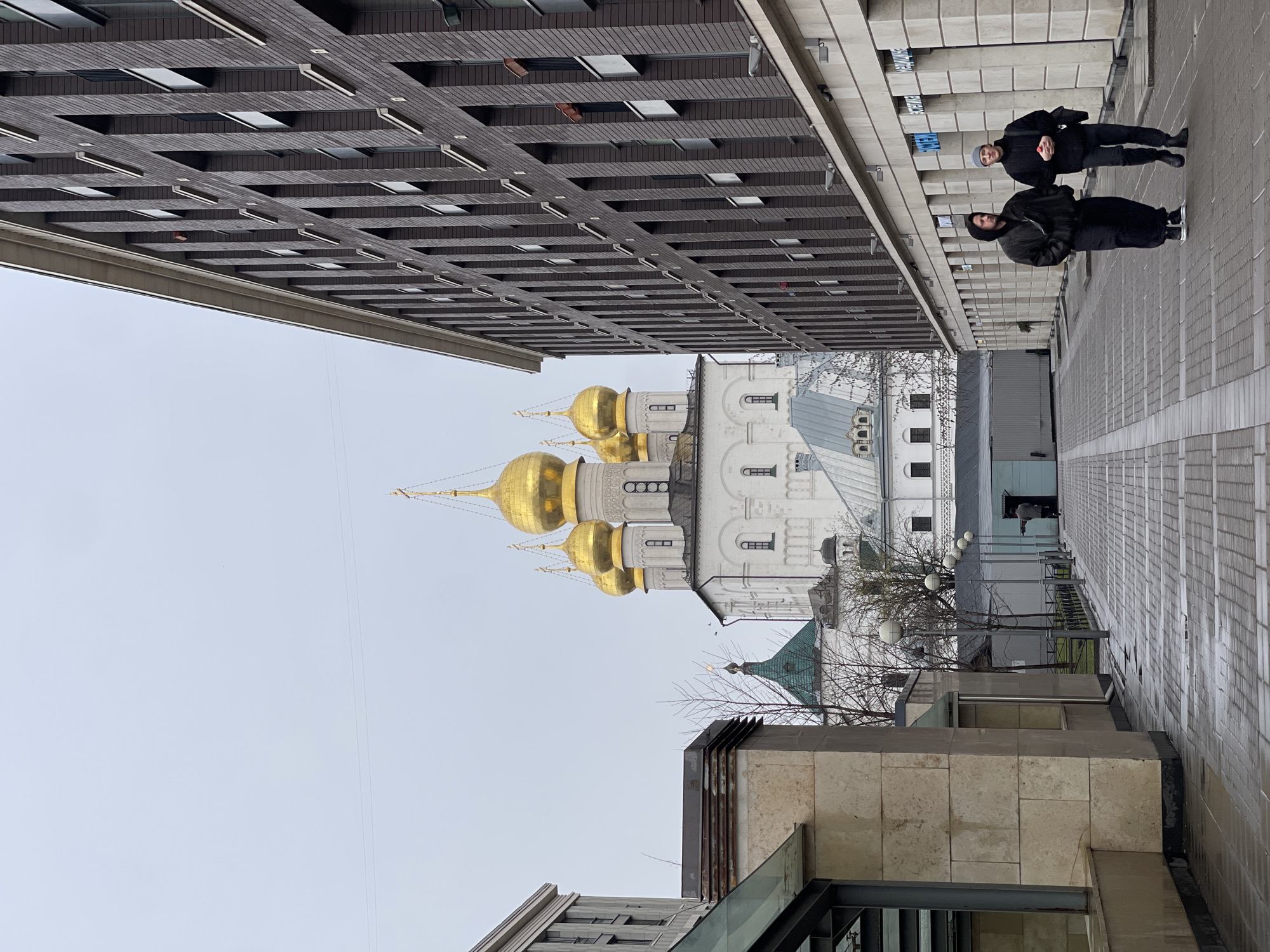 The image shows a view of Saint Petersburg, Russia, angled upwards. In the center, a white building with golden domes is visible. A modern building with a grid-like facade is on the left. On the right, two people dressed in dark clothing are standing on a tiled walkway. The sky is overcast.