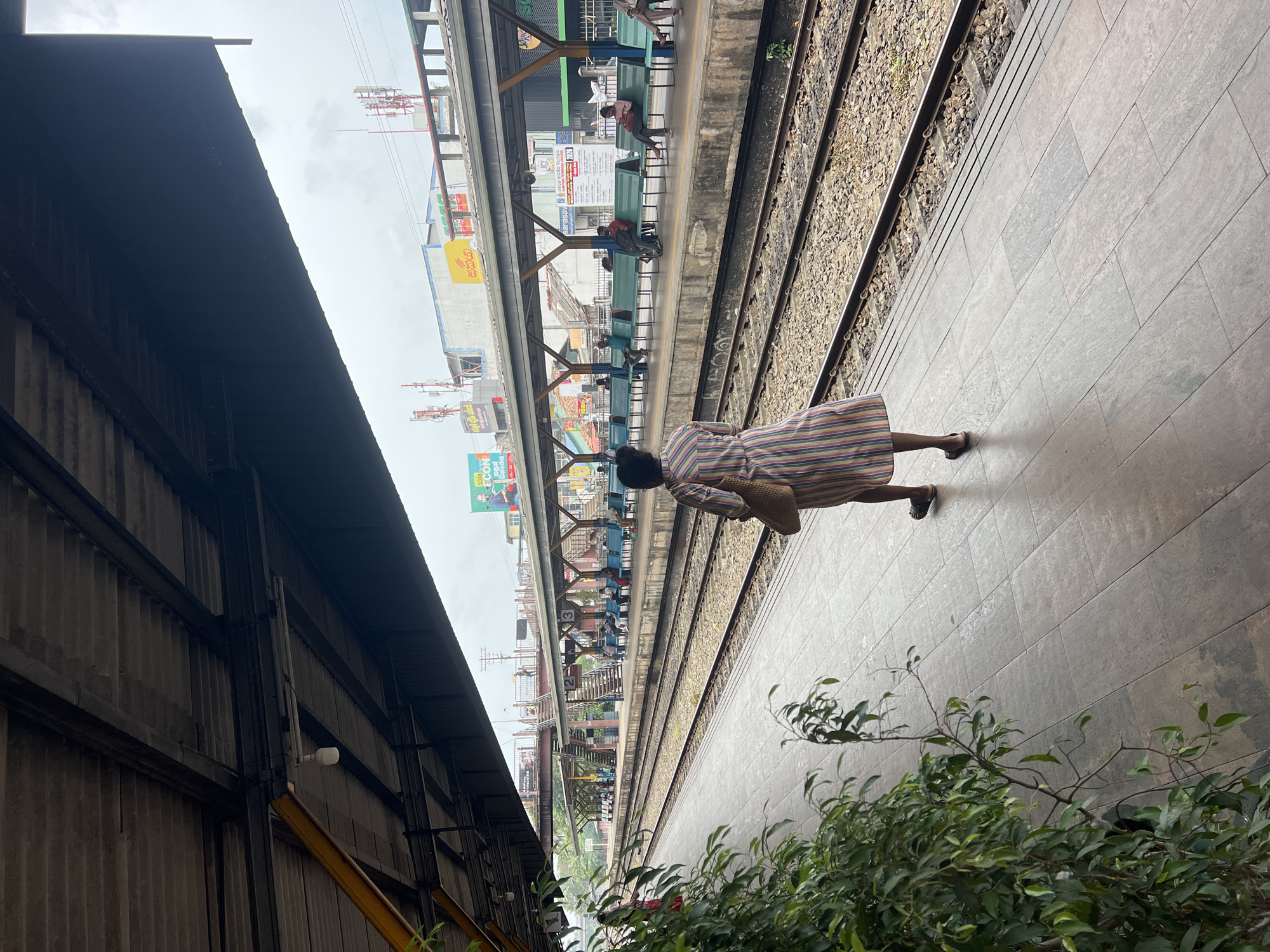 The image depicts a woman standing on a tiled train platform in Gampaha, Sri Lanka. She is wearing a vertically striped dress and carrying a woven bag. The platform is covered by a dark, corrugated roof supported by metal beams, with fluorescent lighting visible. Adjacent to the platform are train tracks, with gravel ballast. Across the tracks, another platform with blue benches is visible, occupied by several people waiting. Beyond this platform, buildings with various signs and advertisements can be seen, along with telecommunication towers on the horizon. Lush green foliage is present on the left side of the foreground. The sky is overcast. The scene appears to be a functioning train station, possibly during daylight hours.