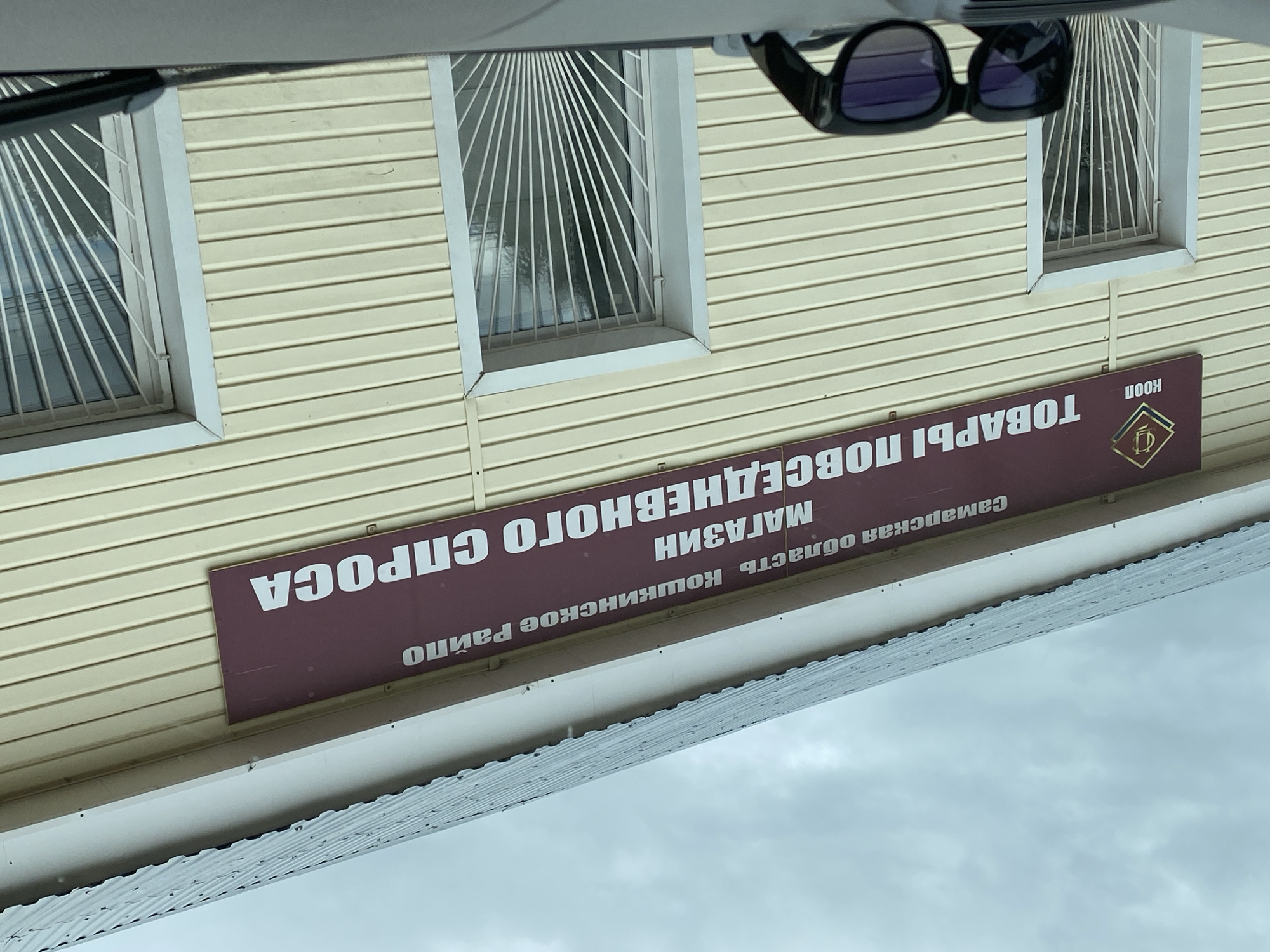 The image shows the facade of a building with a sign in Russian. The sign reads "Самарская область Кошкинское Райпо МАГАЗИН ТОВАРЫ ПОВСЕДНЕВНОГО СПРОСА". This translates to "Samara Region Koshkinsky District Consumer Society STORE OF EVERYDAY GOODS". Below the sign, there are two windows with decorative metal grilles and a logo with the letters "КООП". In the foreground, a pair of dark sunglasses rests on a surface. The building is clad in yellow siding and has a grey metal roof. The sky is overcast. The location is identified as Nadezhdino, Russia.