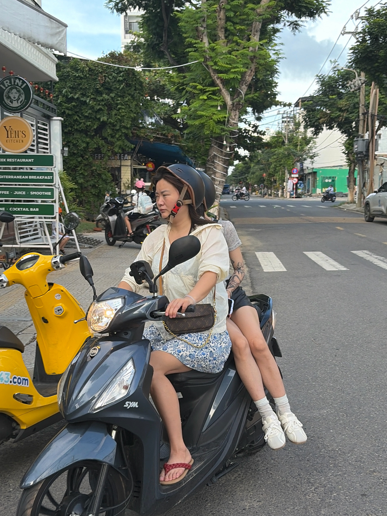 Two individuals, a driver and a passenger, are riding a dark gray Sym scooter on a city street in Đà Nẵng, Vietnam. The driver, a woman wearing a white hooded jacket, a blue and white floral skirt, and a black helmet, is in the foreground. The passenger, wearing a gray t-shirt, is seated behind her. The scooter has its headlights on, and it is traveling on a paved road with white pedestrian crossing lines. In the background, there are trees, buildings, utility poles, and other vehicles, including a yellow scooter to the left of the main subject. Signs for "IRINI" and "YEN'S KITCHEN" are visible on a building to the left of the frame. The sky is overcast.