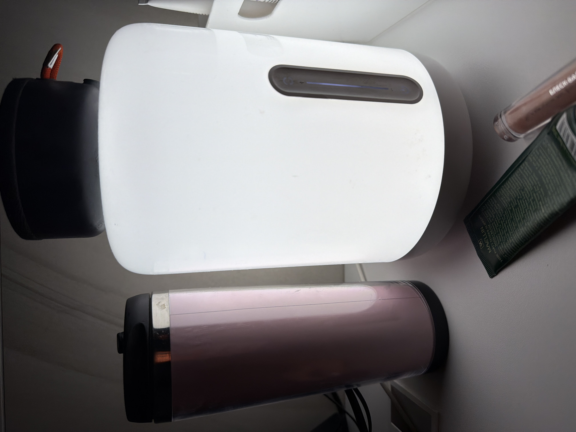 The image depicts a close-up, angled view of several items on a white surface, likely a bathroom counter.
A white, oblong object with a dark gray rectangular slot on top dominates the center. To the left is a black cylinder with a small orange accent. Below the white object is a pink cylindrical object with black end caps and silver accents. To the right, partially overlapping, are a tube of green cream and a lipstick tube. The items appear to be toiletries or personal care products. The scene is artificially lit, casting soft shadows.
There is no information to infer that the image was taken in Saint Petersburg, Russia.
