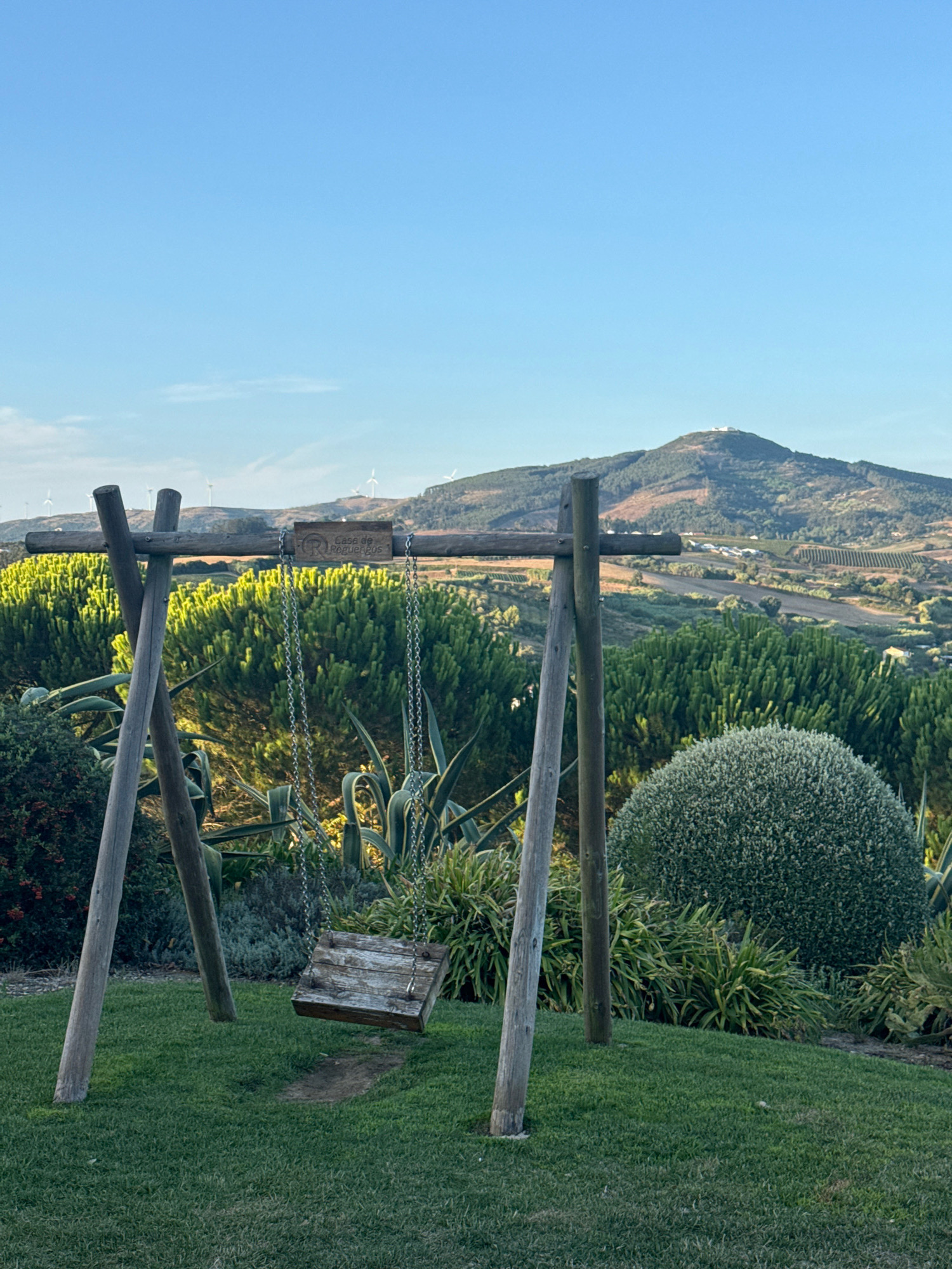 A wooden swing set with a rustic wooden seat hangs from metal chains, suspended from a weathered wooden frame. The swing is positioned on a well-maintained green lawn. In the background, a lush landscape unfolds, featuring dense green shrubs, agave plants, and a large, perfectly round, manicured green bush. Beyond this vibrant vegetation, rolling hills are visible, dotted with vineyards and scattered buildings, suggesting an agricultural or rural setting. The distant horizon is marked by a prominent mountain peak topped with a structure, possibly a building or monument, and several wind turbines are scattered across the undulating terrain. The sky above is a clear, bright blue. The scene appears to be set in the municipality of Mafra, Portugal, likely at an outdoor venue or estate given the manicured surroundings and the presence of a sign on the swing frame reading "Casa de Pegueros." The lighting suggests it is either early morning or late afternoon.