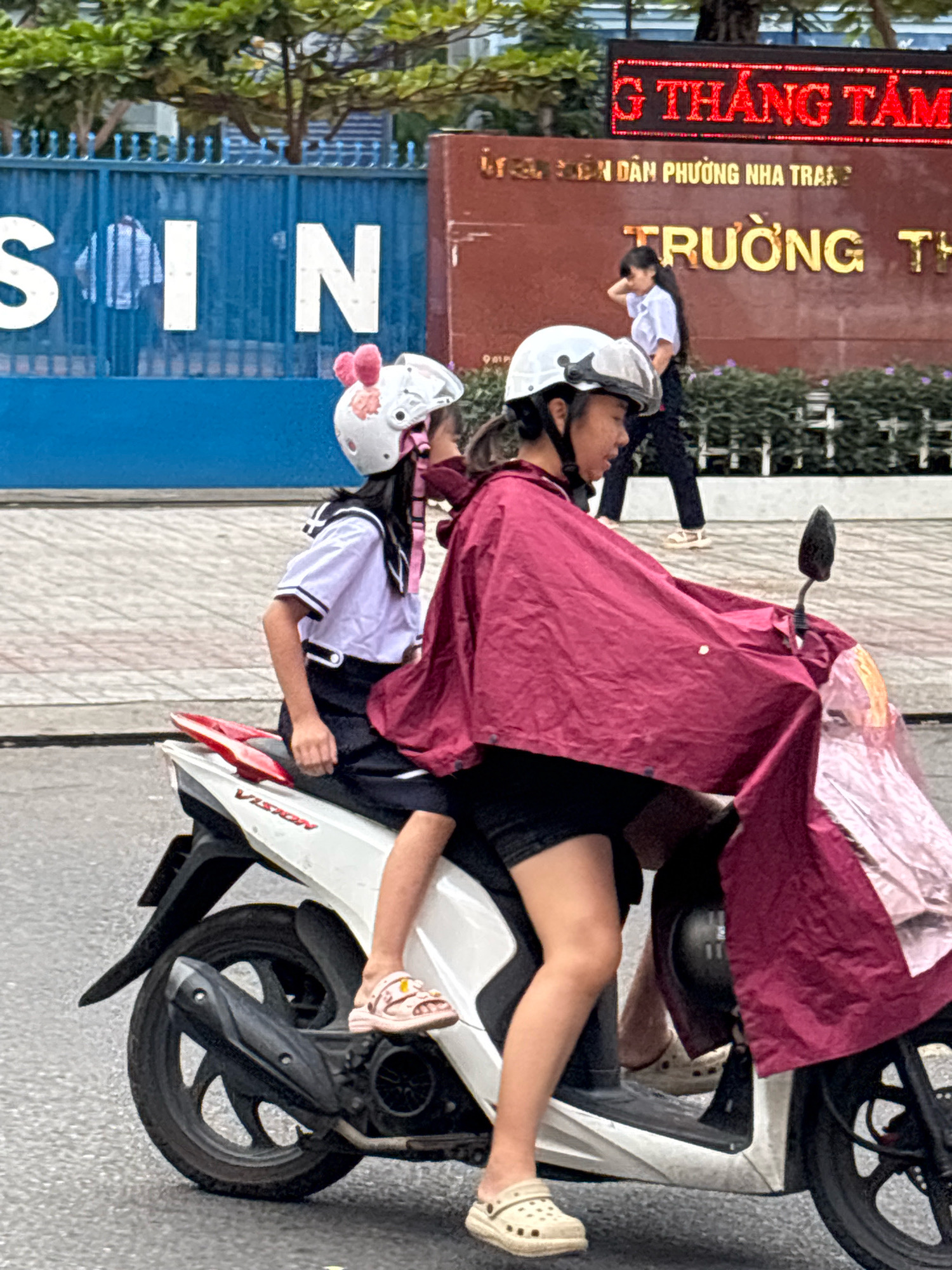In Nha Trang, Vietnam, two young girls ride on a motorcycle. The girl in front, driving, wears a dark shirt and shorts, a white helmet, and a maroon rain poncho that drapes over her and her passenger. The girl behind her, dressed in a sailor-style uniform with a white top and dark skirt, also wears a white helmet adorned with pink bunny ears. Her feet are clad in pink sandals. Both girls are wearing helmets, suggesting safety precautions are being taken.
The scene appears to be a street in Nha Trang, with a blue fence featuring large white letters spelling "SIN" in the background. Beyond the fence, a building with a prominent sign written in Vietnamese characters is visible. The sign features red LED lettering at the top and yellow lettering below, indicating "TRUONG TH" and "G THANG TAM". The girls are on a white motorcycle, with its rear wheel and exhaust pipe clearly visible. The younger girl's sandals are pink and decorated with small yellow charms. The older girl is wearing light beige Crocs. The overall atmosphere seems to be daytime, likely during school hours given the uniforms and the school-related signage in the background. The weather appears to be overcast, as the girl in front is wearing a rain poncho.