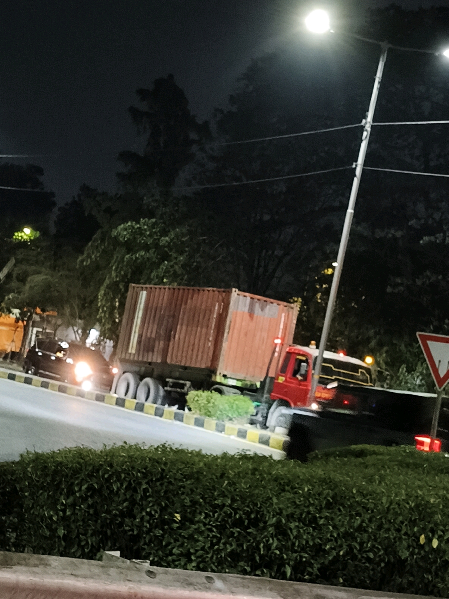 At night in Surabaya, Indonesia, a red truck carrying a large, rusty shipping container is parked on the side of a road. The truck has several wheels visible, and its headlights are on. In front of the truck, a black car with its headlights illuminated is also parked. The road is bordered by a black and yellow striped curb. Behind the truck and car, lush green trees and foliage form a dense background. A street lamp with a bright light is visible in the upper right portion of the image. Further down the road, a triangular traffic sign with a red border is posted. The scene suggests a temporary stop or parking situation on a well-lit street in an urban area.
