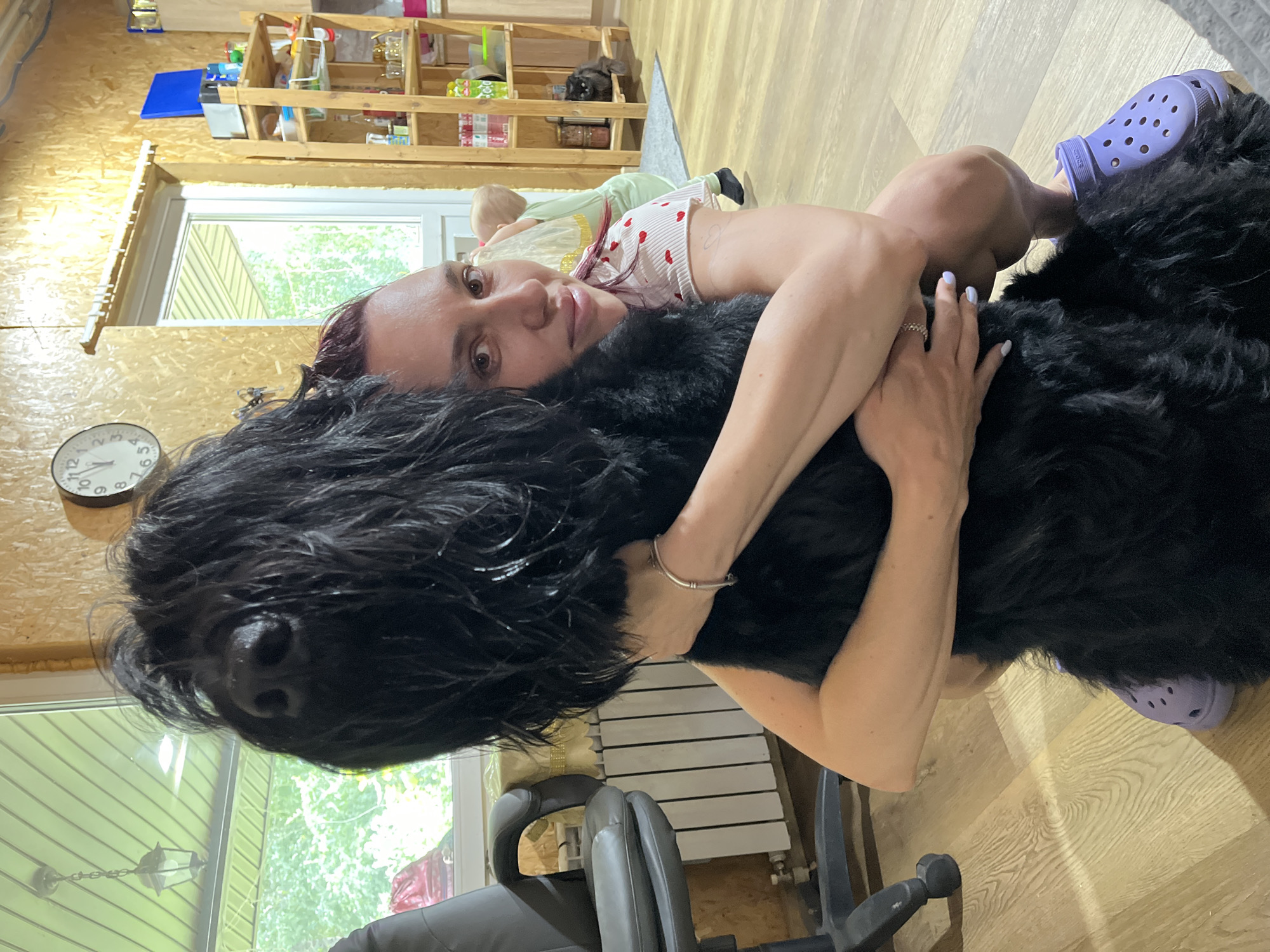 A woman with red hair and a pink and white heart-patterned shirt is hugging a large black dog. She is kneeling on a wooden floor and wearing purple Crocs. A black cat is visible on a wooden shelf in the background, which appears to be a kitchen or pantry area. A child in a light green outfit is also present in the background. The room has wood-paneled walls and a window with a view of greenery. A clock hangs on the wall. The location is identified as Nagorny, Russia.