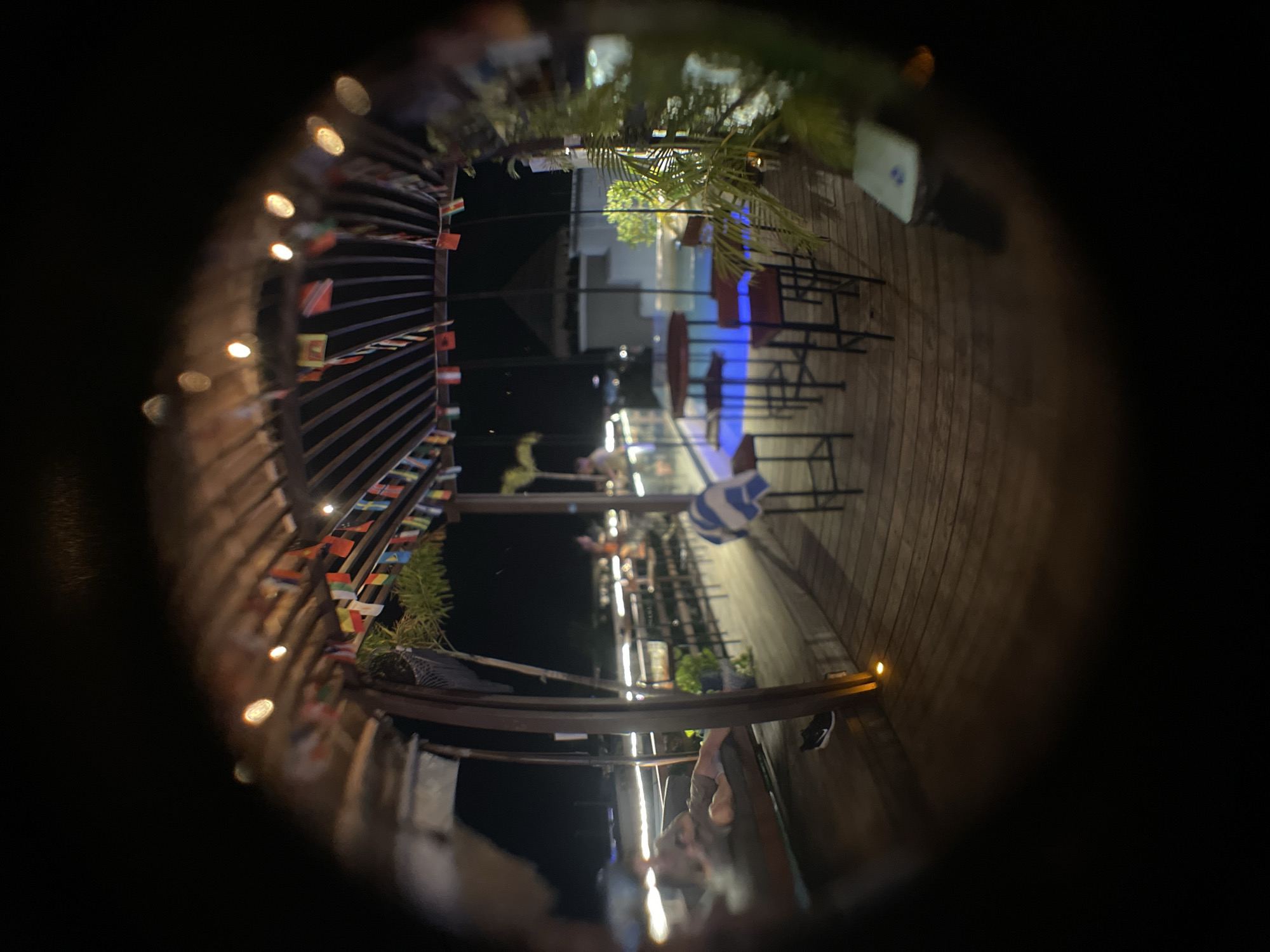 The image, taken in Playa del Carmen, Mexico, presents a nighttime view through a small aperture, possibly a peephole or narrow lens. The scene depicts an outdoor deck or patio area, identifiable by wooden plank flooring and metal railings adorned with numerous small flags, potentially representing different countries. Strings of small lights illuminate the railings. In the background, there's a glimpse of an open-air bar or restaurant area with tables and chairs, possibly with blue-colored lighting. A few plants or palm trees are also visible. At the very bottom of the image, parts of people sitting appear.