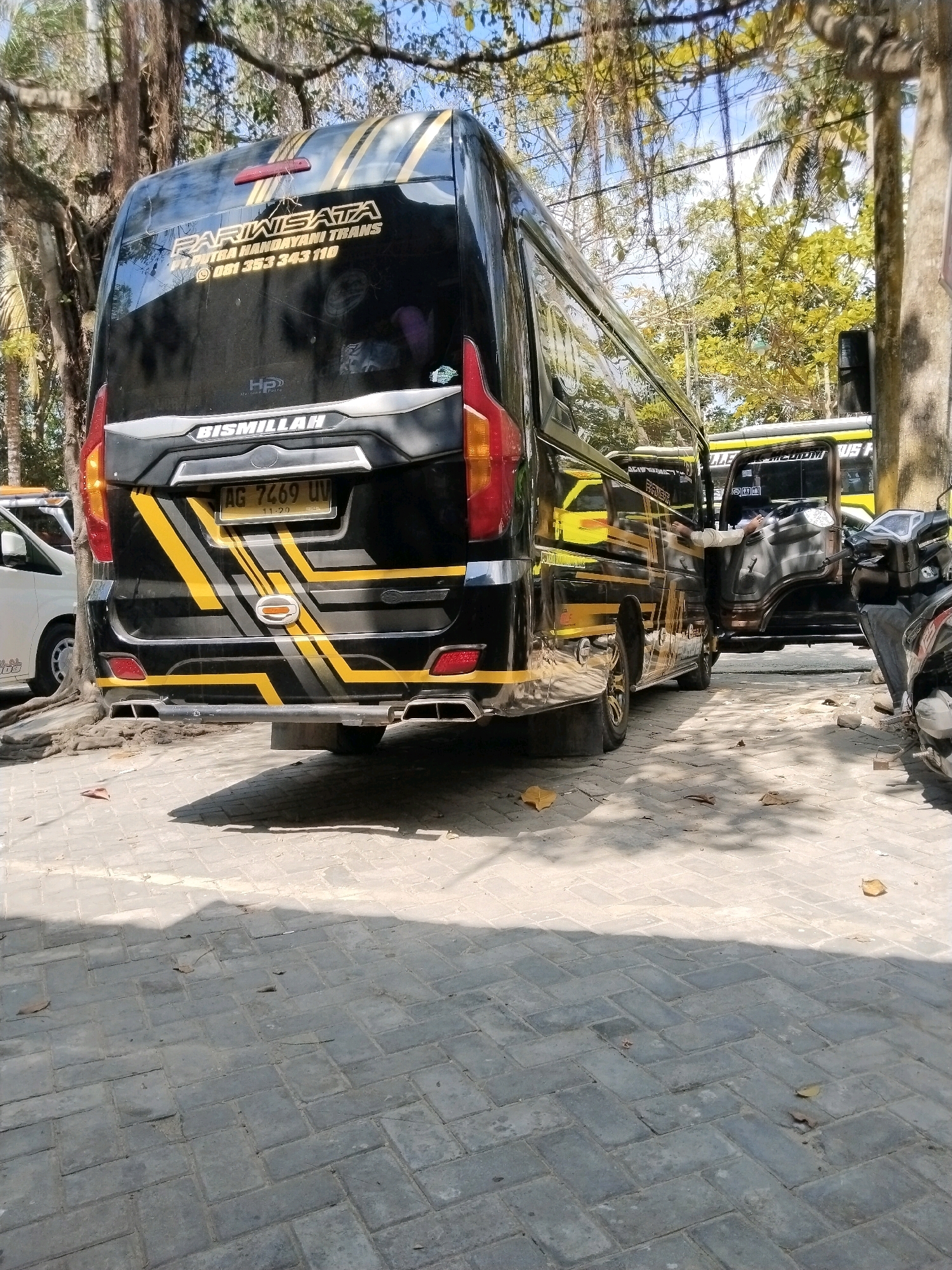This image captures a street scene in Prigi, Indonesia, focusing on the rear of a black and yellow tour bus. The bus, labeled "PARIWISATA" and "PUTRA HANDAYANI TRANS", displays a license plate reading "AG 7469 UV". To its right, another yellow bus with open doors reveals a person sitting inside. A parked motorcycle is visible in the foreground on the right. The ground is paved with interlocking gray bricks, and fallen leaves are scattered across the surface. Lush green trees with hanging vines provide a natural backdrop, and the sky is partially visible through the foliage. The lighting suggests it is daytime, with shadows cast by the trees and vehicles.