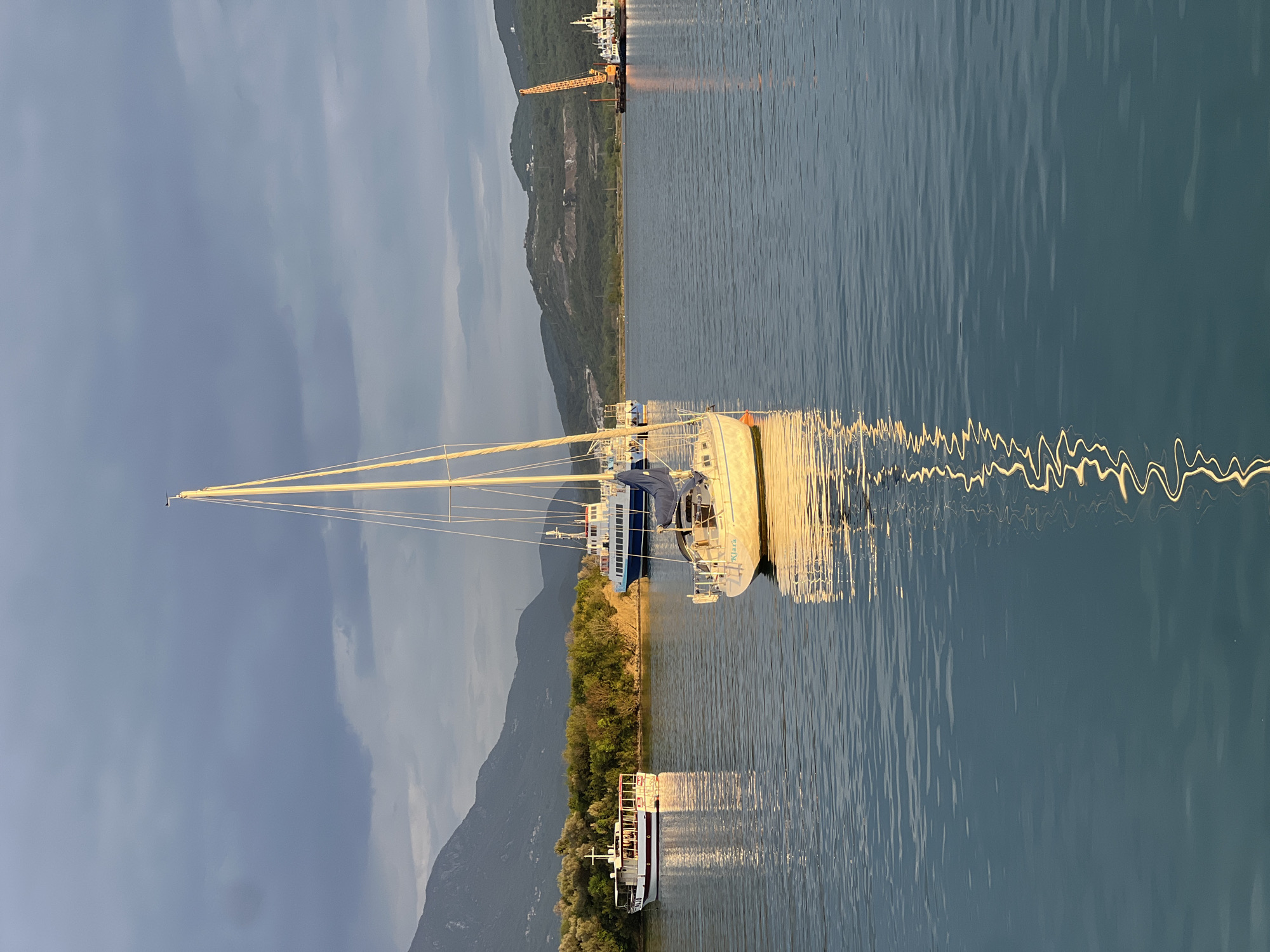 The image depicts a waterfront scene in Đuraševići, Montenegro, featuring a sailboat docked in the water. The sailboat is white with a tall mast and blue accents. It is located near a shoreline with green vegetation. In the background, mountains rise into a cloudy sky. The water surface is reflective, displaying the outlines of the boat and nearby objects. A construction crane is visible on the distant shore, suggesting a harbor or shipyard setting. Another boat, appears to be a tour boat, is also docked along the shoreline. The scene is illuminated by a soft, indirect light, possibly from the late afternoon sun.