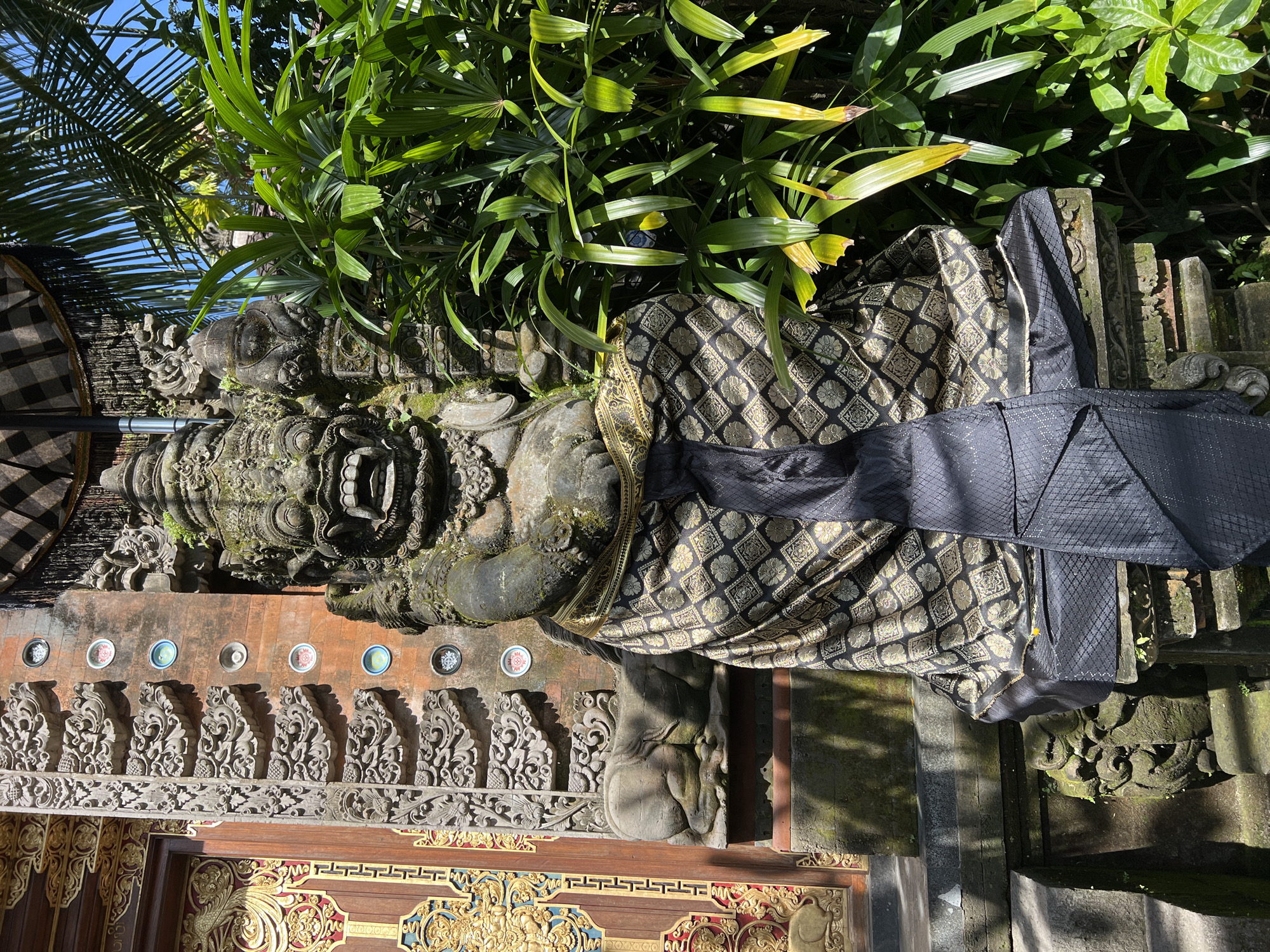 A stone statue in Petulu, Indonesia, adorned with a patterned cloth and a black sash, stands partially obscured by lush green plants. The statue's intricate details are weathered with age. Behind the statue is a decorative wall with a row of circular ornaments and ornate carvings along the top, above a wooden door with golden embellishments. A woven umbrella is visible on the left side of the statue. The scene is bathed in sunlight, creating shadows that add depth to the stone and foliage.