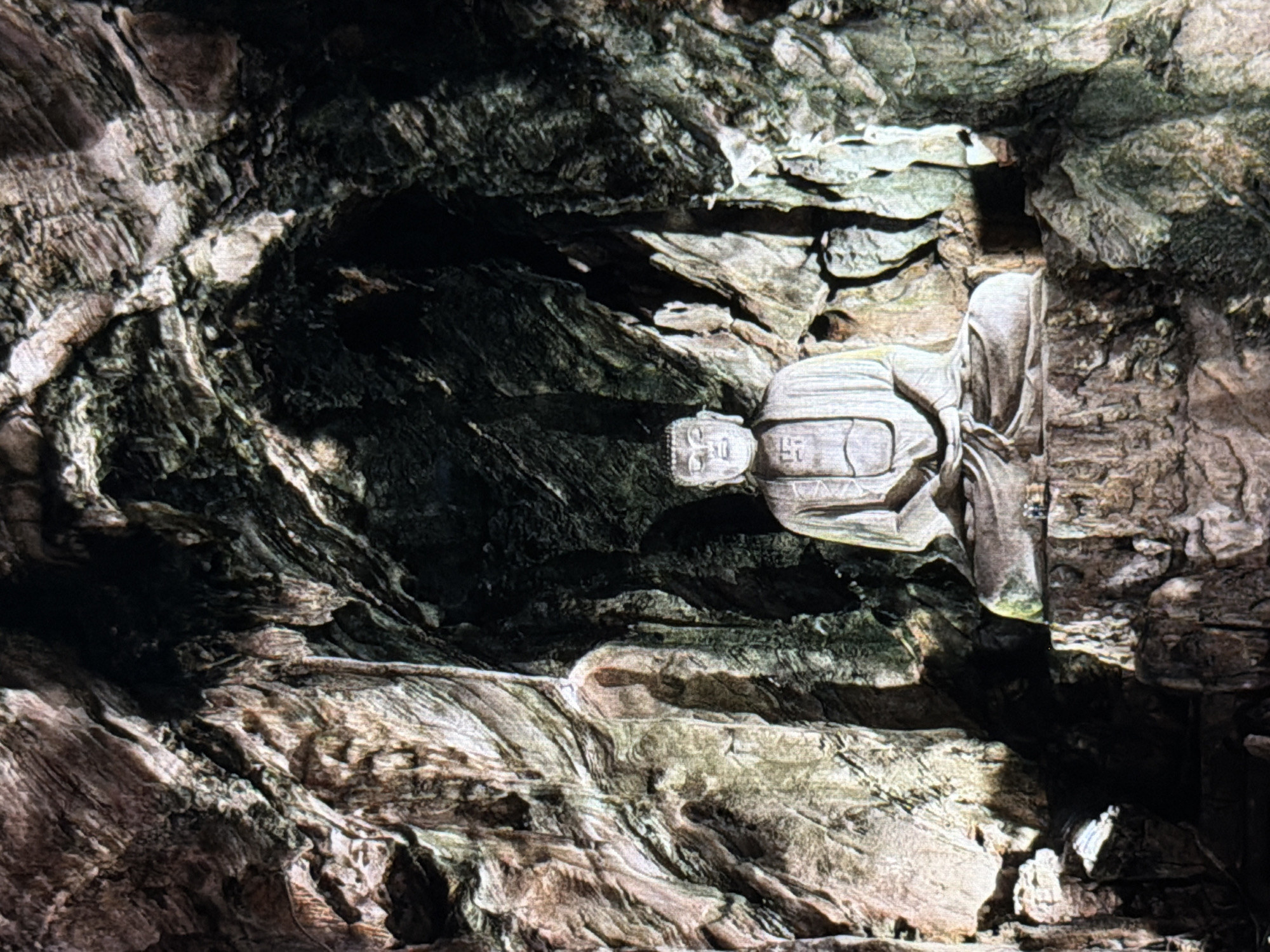 A stone statue of a figure, potentially a Buddha or monk, is embedded within a rocky cave-like structure. The statue is carved in a stylized, blocky fashion and appears to be made of a similar stone to the surrounding environment. The head of the figure features inscribed symbols. The cave is formed by dark and light-colored rocks with layered patterns. This is likely located within the Marble Mountains in Đà Nẵng, Vietnam, known for their cave temples and stone sculptures.