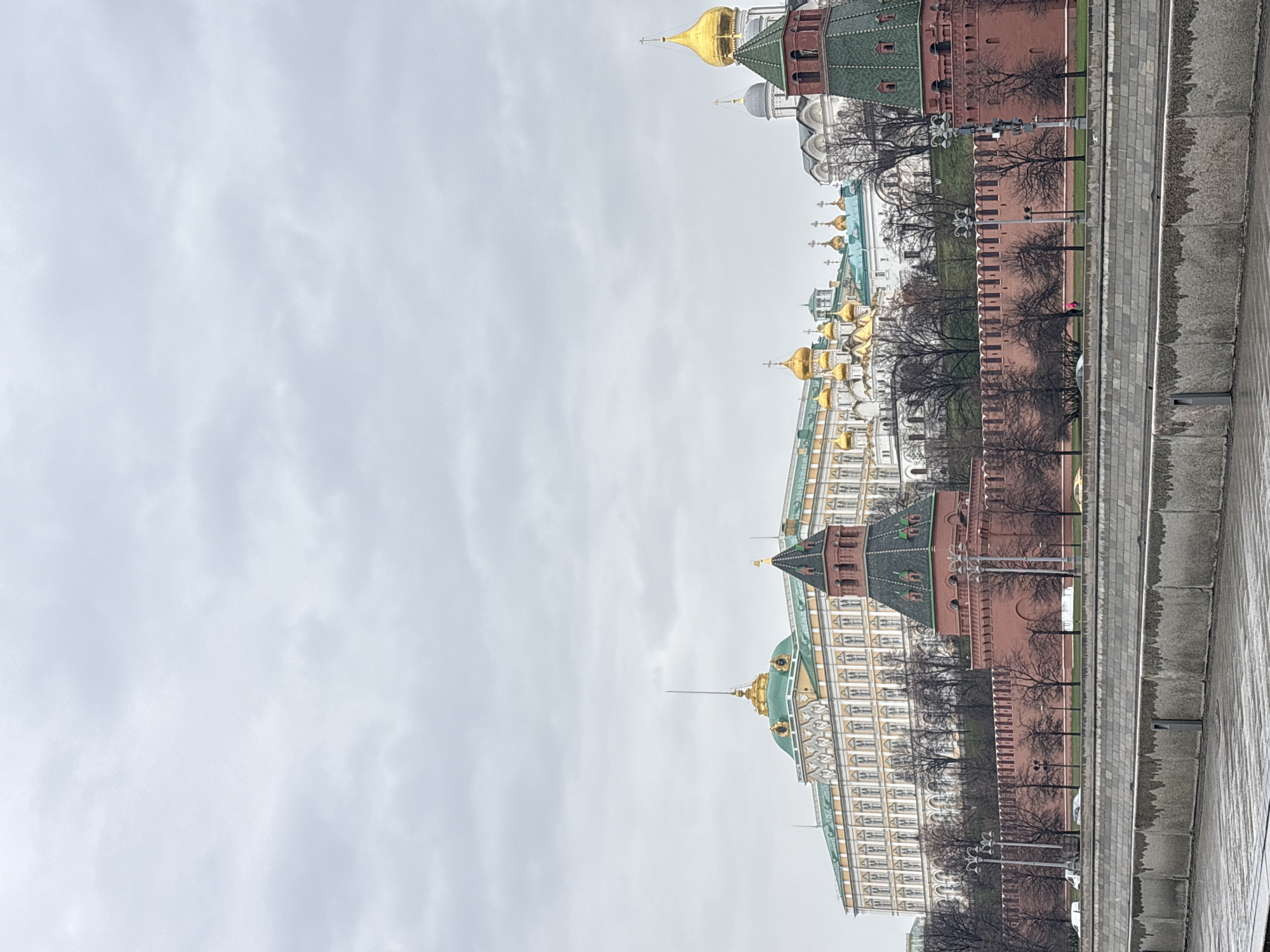 The image depicts the Moscow Kremlin complex under a cloudy sky. Visible within the complex are walls, towers, golden domes of cathedrals, and other architectural details. The composition suggests the view is taken from a vantage point across a waterway or street from the Kremlin. The surrounding environment includes leafless trees bordering the embankment. The sky is overcast, indicating cool weather conditions. No people are clearly visible in the immediate foreground.