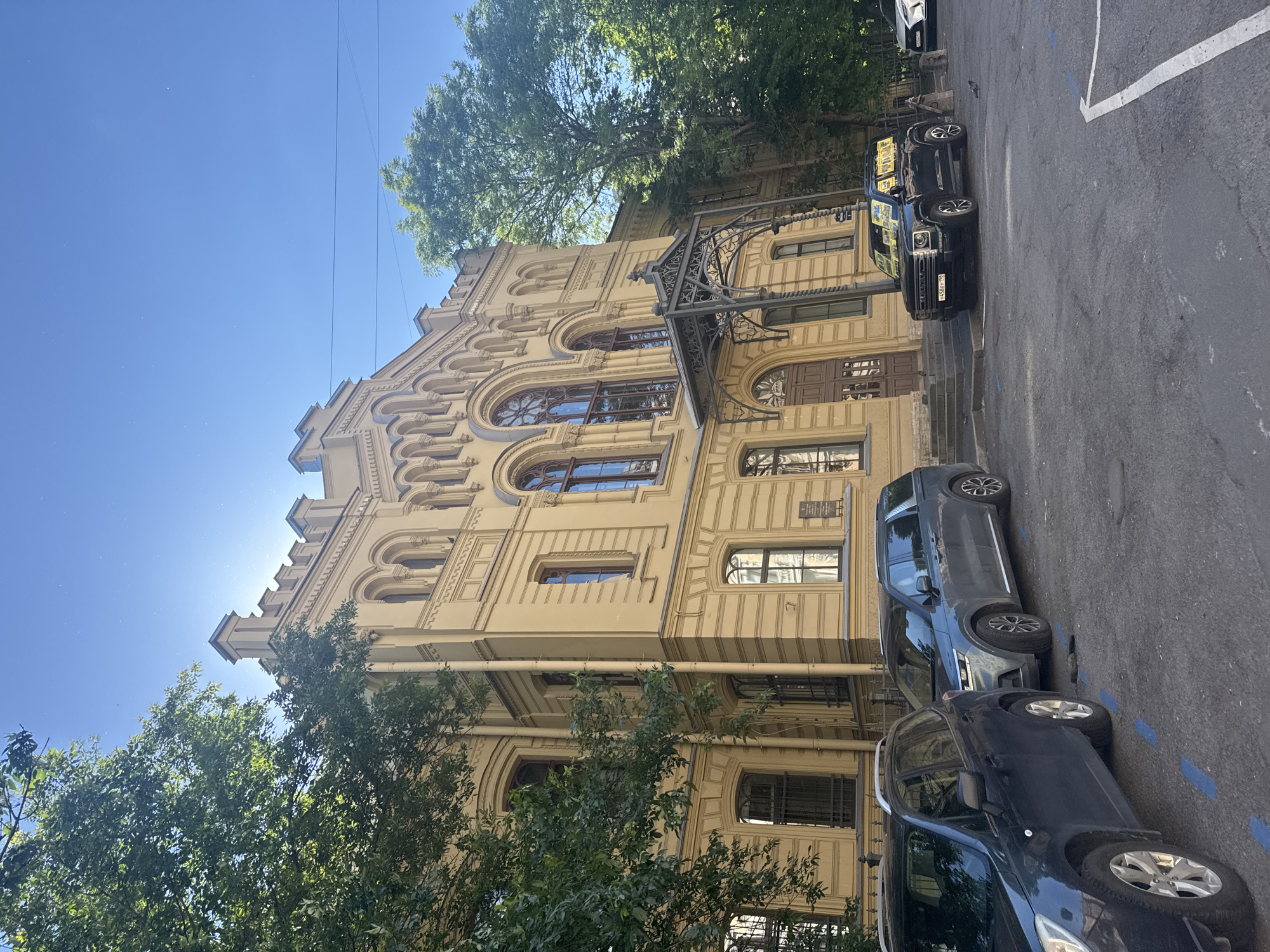 The image depicts the facade of the yellow, neo-Gothic building of the Russian Museum of Ethnography, located at 4, Inzhenernaya Street, in Saint Petersburg, Russia. The building features arched windows, decorative battlements, and a wrought-iron canopy over the entrance. Several cars are parked in front of the building, including a dark blue SUV on the left, a gray minivan in the middle, and a black SUV on the right. Trees with lush green foliage flank the building, and the sky is clear and blue with a few visible dust particles. The scene is captured in daylight under a bright sun.
