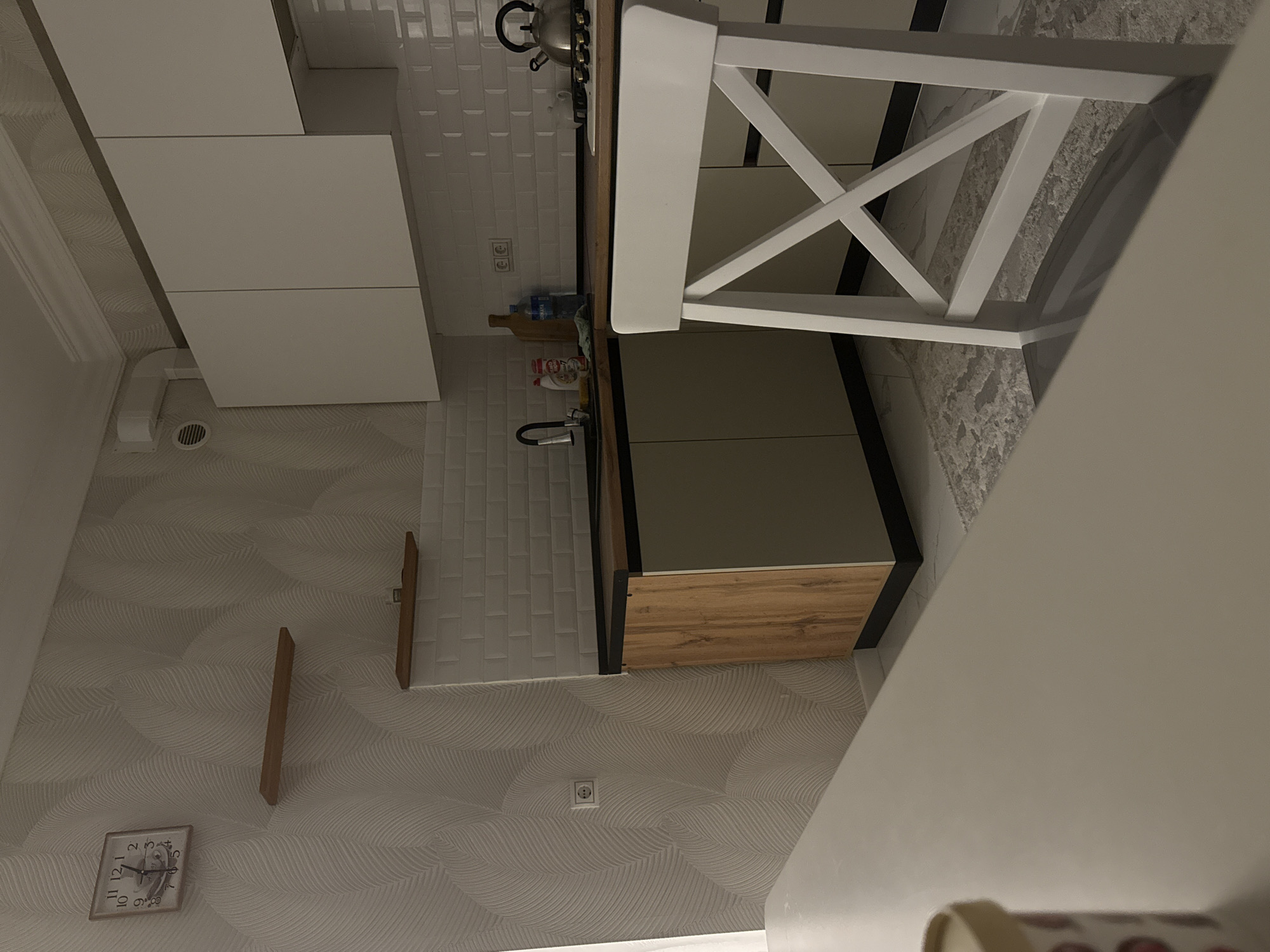 The image depicts a modern kitchen with a light gray, textured wallpaper and white subway tile backsplash. A white chair with a distinctive cross-back design is positioned at a wooden-topped counter. Below the counter, there are light gray cabinet doors and a wooden drawer front, creating a contemporary aesthetic. On the counter, a silver kettle and a few bottles are visible.
To the left of the counter, white cabinets are mounted on the wall, with a ventilation duct visible below them. The wall also features a square clock with Roman numerals. Further to the left, two narrow wooden shelves are mounted on the wallpapered wall. On the right side of the image, the wall has a rough texture and a white electrical outlet is present.
The scene appears to be indoors, likely in a residential kitchen. The lighting suggests it could be daytime or evening, with artificial light sources contributing to the illumination. There are no people visible in the image. The overall impression is one of a clean and stylish, yet lived-in, kitchen space. The specific location is Derbent, Russia, but this is not visually evident from the image itself.