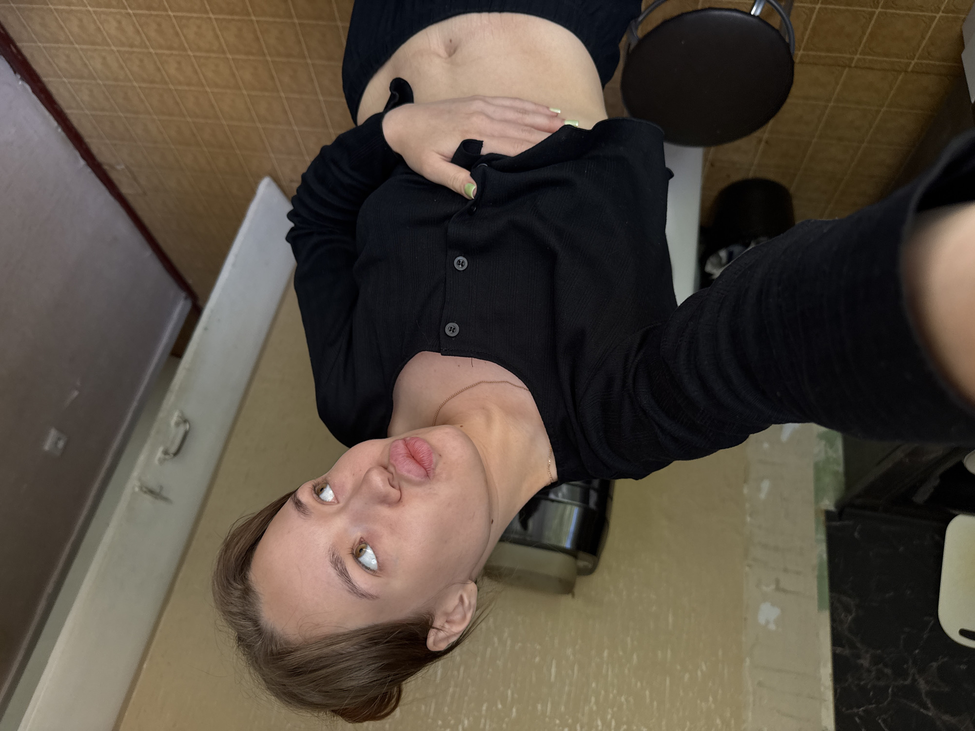 A young woman with light brown hair pulled up in a bun takes a selfie in what appears to be a residential interior. She is wearing a black long-sleeved, cropped cardigan over dark pants and is holding her stomach with her left hand, revealing her midriff. Her facial expression is a playful pout with her eyes looking to the right. The background shows beige walls, a dark countertop with a white object on it to the left, and a brown door or closet to the right. A black stool and a dark cylindrical object are also visible in the room. The floor is tiled with a diamond pattern. The image is taken from a high angle, looking down at the woman. The overall impression is of a casual, personal moment captured in a home setting. There is no visible indication of the location being specifically within the city of Chaikovsky, Russia.