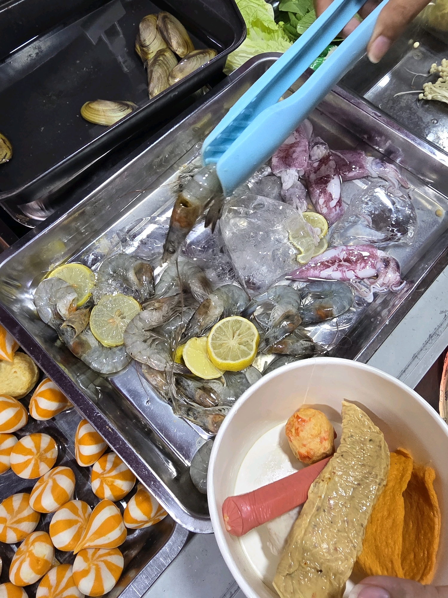 In Tanjung Pinang, Indonesia, a close-up, overhead shot depicts a selection of fresh seafood and processed food items prepared for a meal. In the foreground, a white bowl contains various ingredients: a reddish-pink sausage, a golden-brown, flat piece of processed seafood, and two orange, flat pieces of processed seafood. To the left of the bowl, a tray of striped fish balls, orange and white, is arranged.
The main focus is a large metal tray filled with ice, fresh seafood, and lemon slices. This tray contains numerous raw shrimp and several squid. Blue tongs are actively being used to pick up a shrimp from this tray. Adjacent to this tray, another metal tray holds several clams. In the background, there are glimpses of fresh green lettuce and enoki mushrooms, suggesting a hot pot or similar culinary preparation. The lighting is bright and natural, highlighting the textures and colors of the food. The scene appears to be set in a marketplace or a restaurant specializing in seafood.