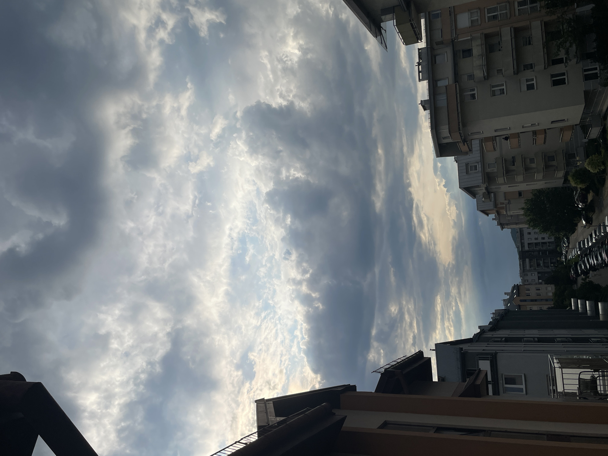 A vertical, high-angle shot captures a dramatic sky filled with a mix of light and dark clouds. The clouds are textured, with sunlight breaking through in some areas, illuminating the undersides of others. In the foreground, the sides of apartment buildings frame the view. The buildings have balconies and windows, with some showing architectural details like slanted roofs. The color palette is dominated by grays and blues from the sky, with warm tones from the building exteriors. The scene suggests an urban environment with a sense of impending weather or the aftermath of a storm.