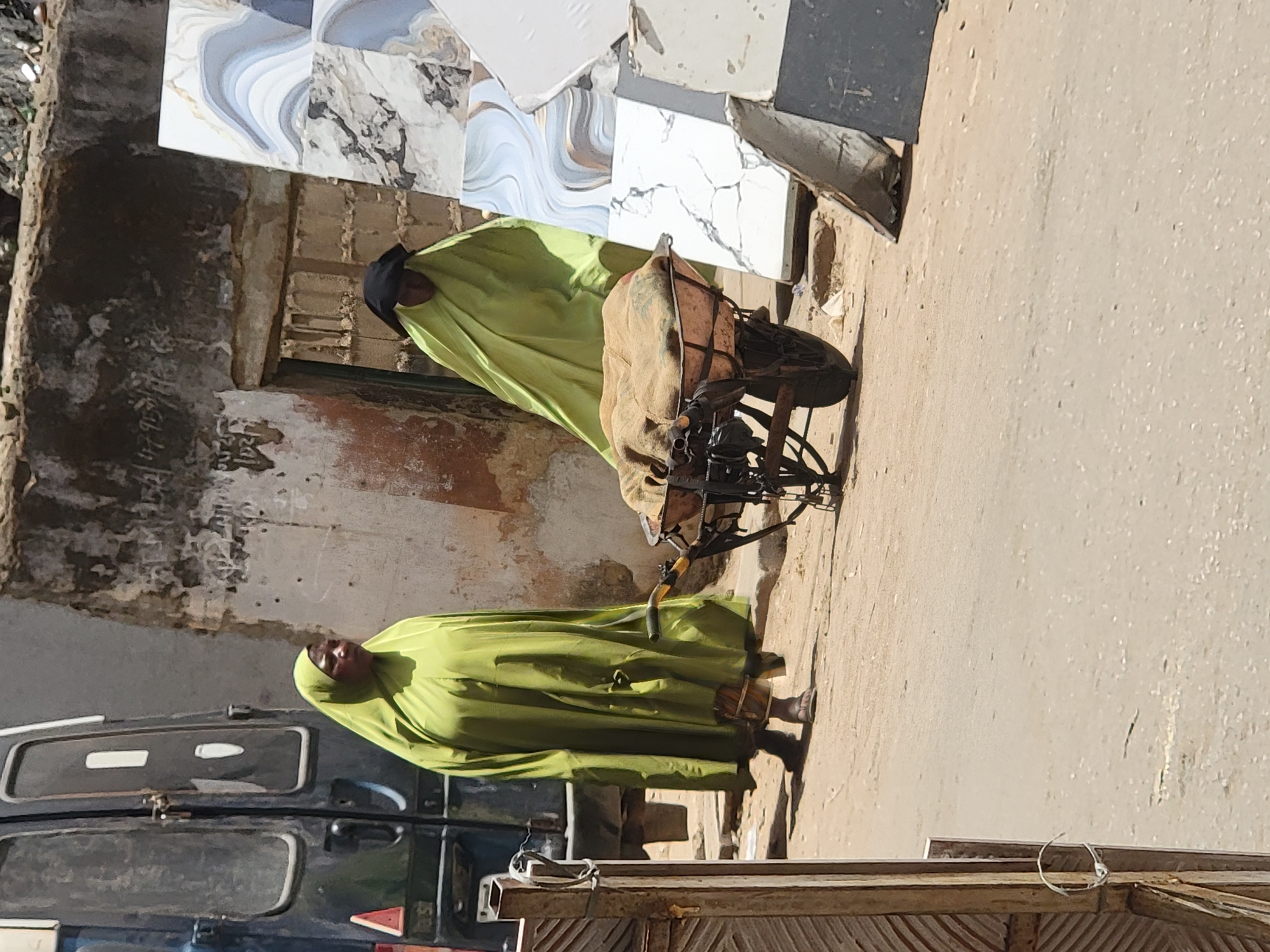 Two people wearing similar green, full-body coverings are visible on a street in what appears to be Maiburiji, Nigeria. One woman stands near the curb in front of a dark-colored van. The other woman stands in front of a cart laden with a covered object or sacks and a bicycle. The background includes walls with deteriorating finishes and discarded or stacked tiles. The street is paved and shows signs of wear. A partially visible wooden structure borders the scene on the right. The lighting indicates a sunny day.