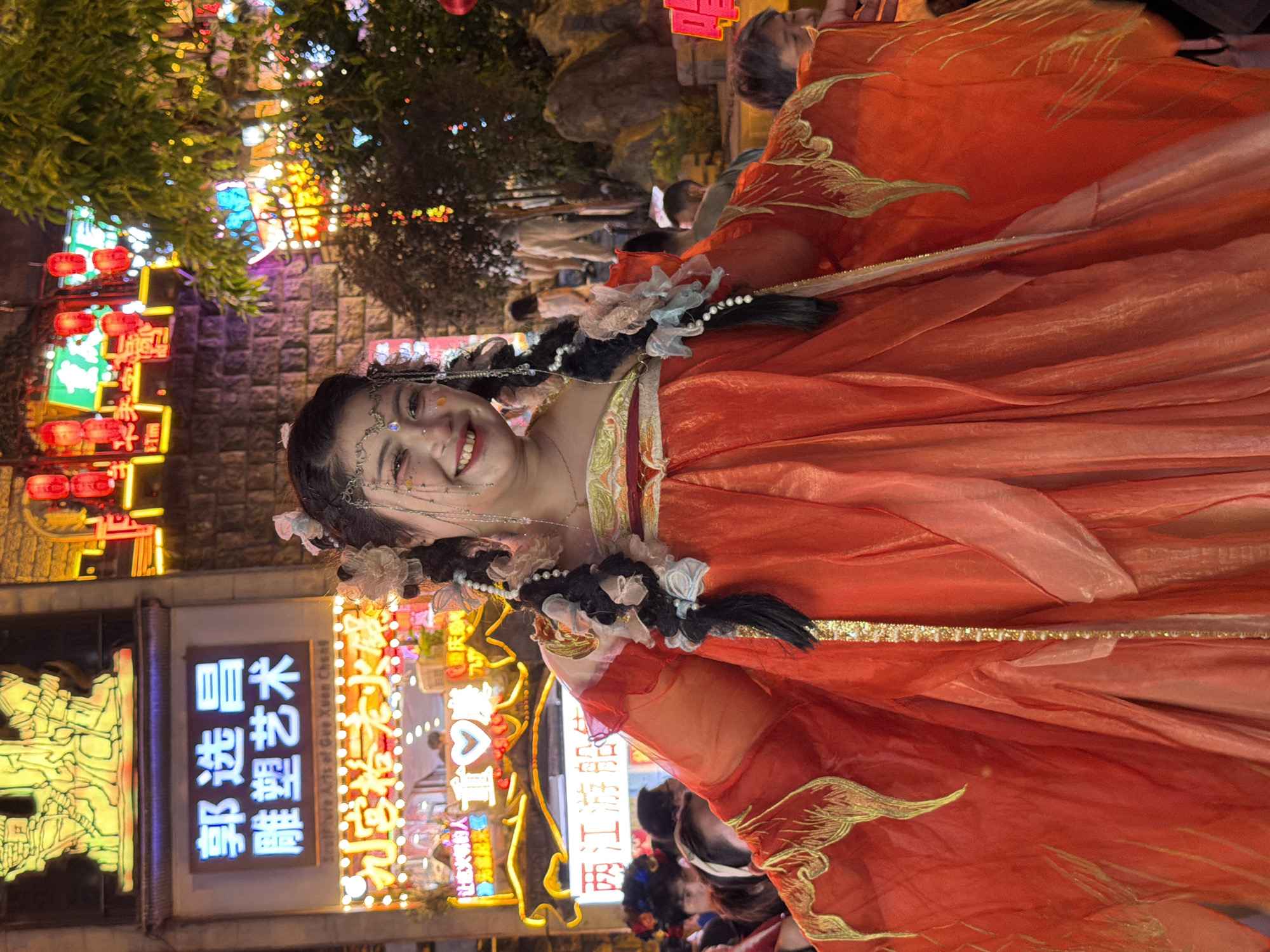 This is a vertical, medium-shot photograph of a young woman in a vibrant orange costume with gold trim and embellishments, reminiscent of traditional Chinese attire. She is adorned with delicate beaded jewelry around her head and neck, and her dark hair is styled in braids decorated with small white flowers and ribbons. She has a bright, friendly smile and glitter makeup on her face. The background suggests a lively night market or street festival in Yuzhong District, China.
Behind the woman, neon signs and bright lights illuminate the scene. On the left, a sign with Chinese characters in yellow neon reads "郭选昌 雕塑艺术" (Guo Xuan Chang Sculpture Art). To the right, another glowing sign features Chinese characters and a heart symbol, with English text below that seems to be part of a business name, possibly related to Chongqing and "two rivers cruise ship." Red lanterns are also visible, adding to the festive atmosphere. People are milling about in the background, indicating a public gathering or event. The overall mood is cheerful and celebratory, capturing a moment of joy amidst the bustling night life of a Chinese city. The lighting suggests it is nighttime.