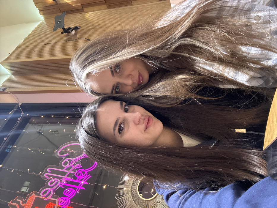 Two young women pose closely. The woman at the top has long blonde hair and is wearing a plaid shirt. The woman in the bottom has long brunette hair and wears a blue jacket. Behind them, there's a window reflecting buildings, and a neon sign that reads "Coffee". The background features a wooden wall with a mounted TV bracket and a decorative mirror.