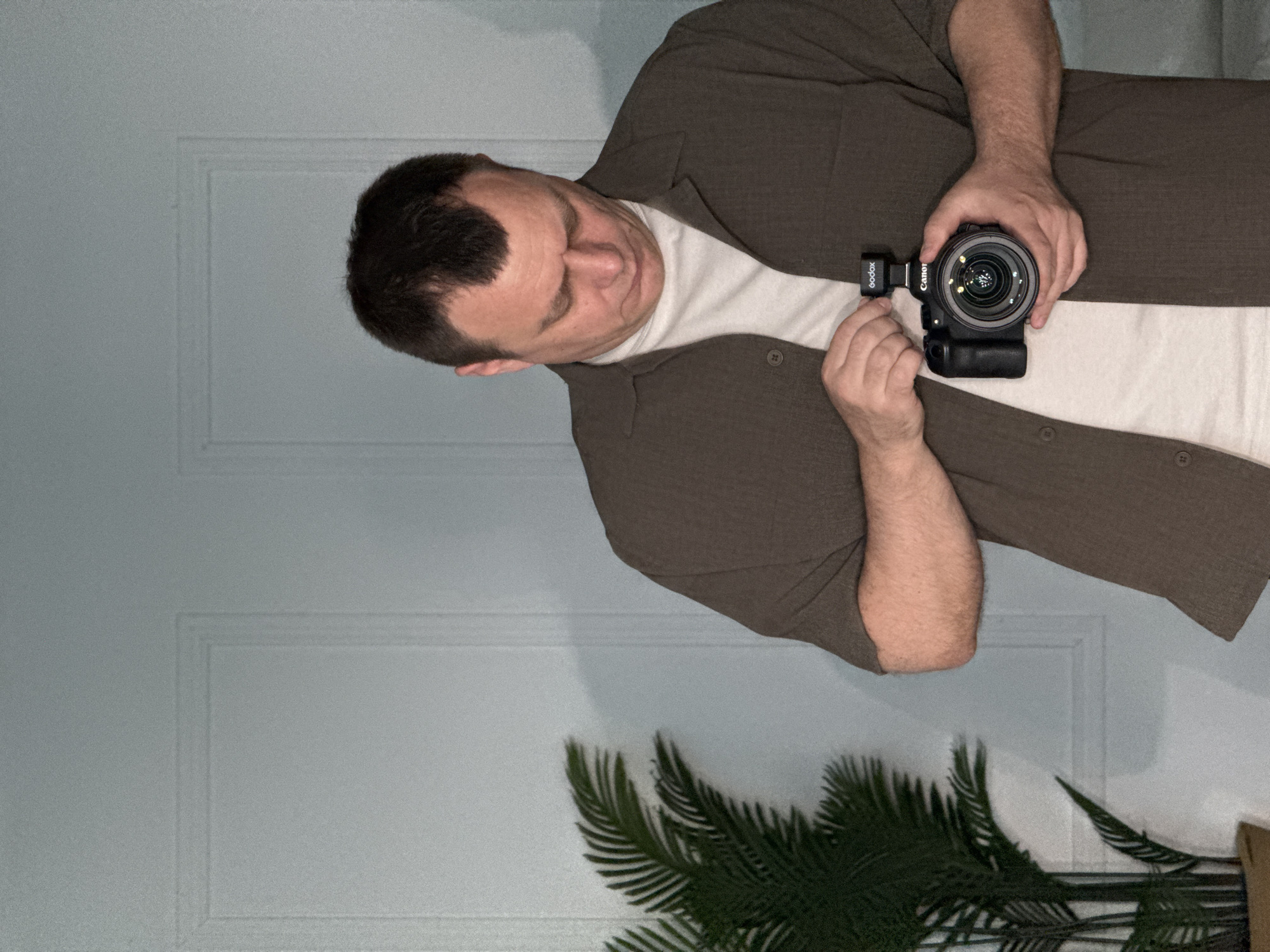 A man is shown holding a Canon camera with a Godox flash attachment. He is wearing a white t-shirt underneath a brown button-down shirt. The background includes a light blue wall with a door frame and a green plant. The location is unknown and there is no indication of being in Chaikovsky, Russia.