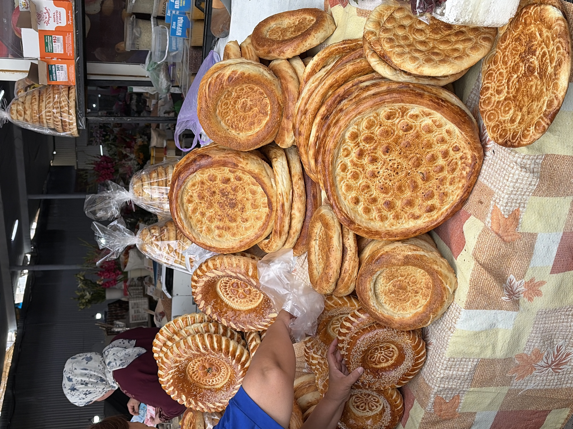 The image shows a close-up view of a market stall overflowing with various types of flatbread. The breads are circular and golden-brown, with some featuring intricate patterns pressed into their surfaces, and others sprinkled with sesame seeds. They are stacked in piles, some are wrapped in clear plastic. In the background, a woman wearing a headscarf is visible, seemingly attending to the stall. The stall is covered with a checkered tablecloth featuring floral patterns. The overall impression is a vibrant display of freshly baked goods at a bustling market.