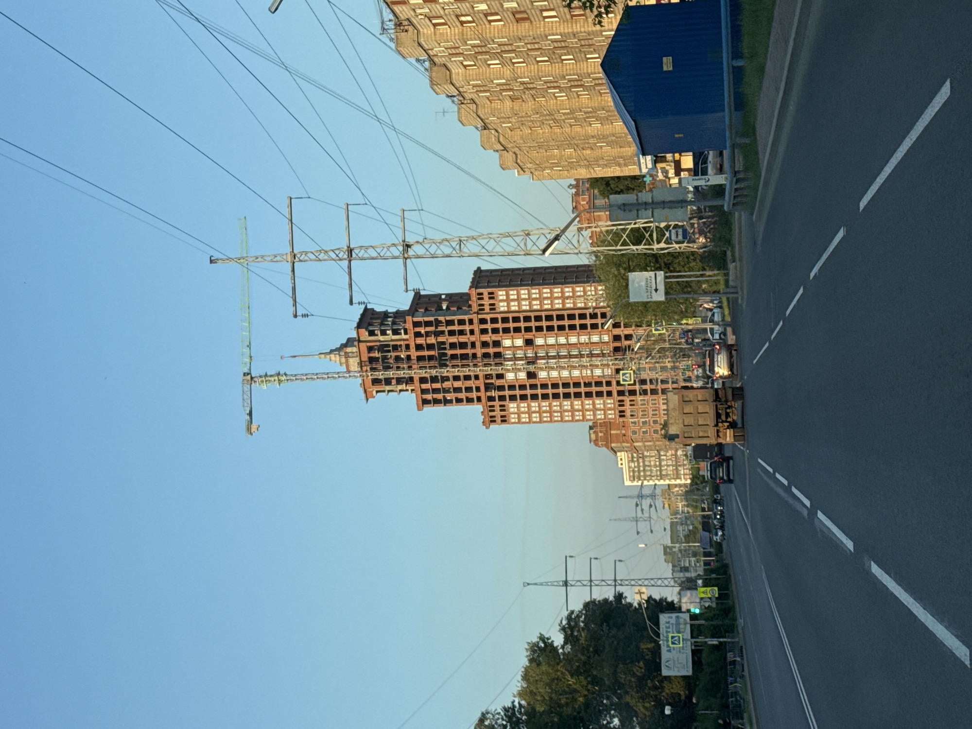 The image depicts an urban scene with a focus on a tall, unfinished skyscraper under construction. The skyscraper, made of red brick and concrete, has a complex structure with visible scaffolding and cranes at its upper levels. It is surrounded by other apartment buildings, some of which appear to be completed and residential, characterized by their repetitive window patterns. Power lines and tall transmission towers are prominent features of the landscape, stretching across the sky and near the buildings. A road with multiple lanes runs through the foreground, with several vehicles, including cars and a truck, driving on it. The environment suggests a sunny day with a clear blue sky. The presence of construction and residential buildings, along with transportation infrastructure, indicates a developing urban area. The signs in Russian suggest this location is in Russia. Further research would be needed to pinpoint the exact location within Tomsk, Russia.