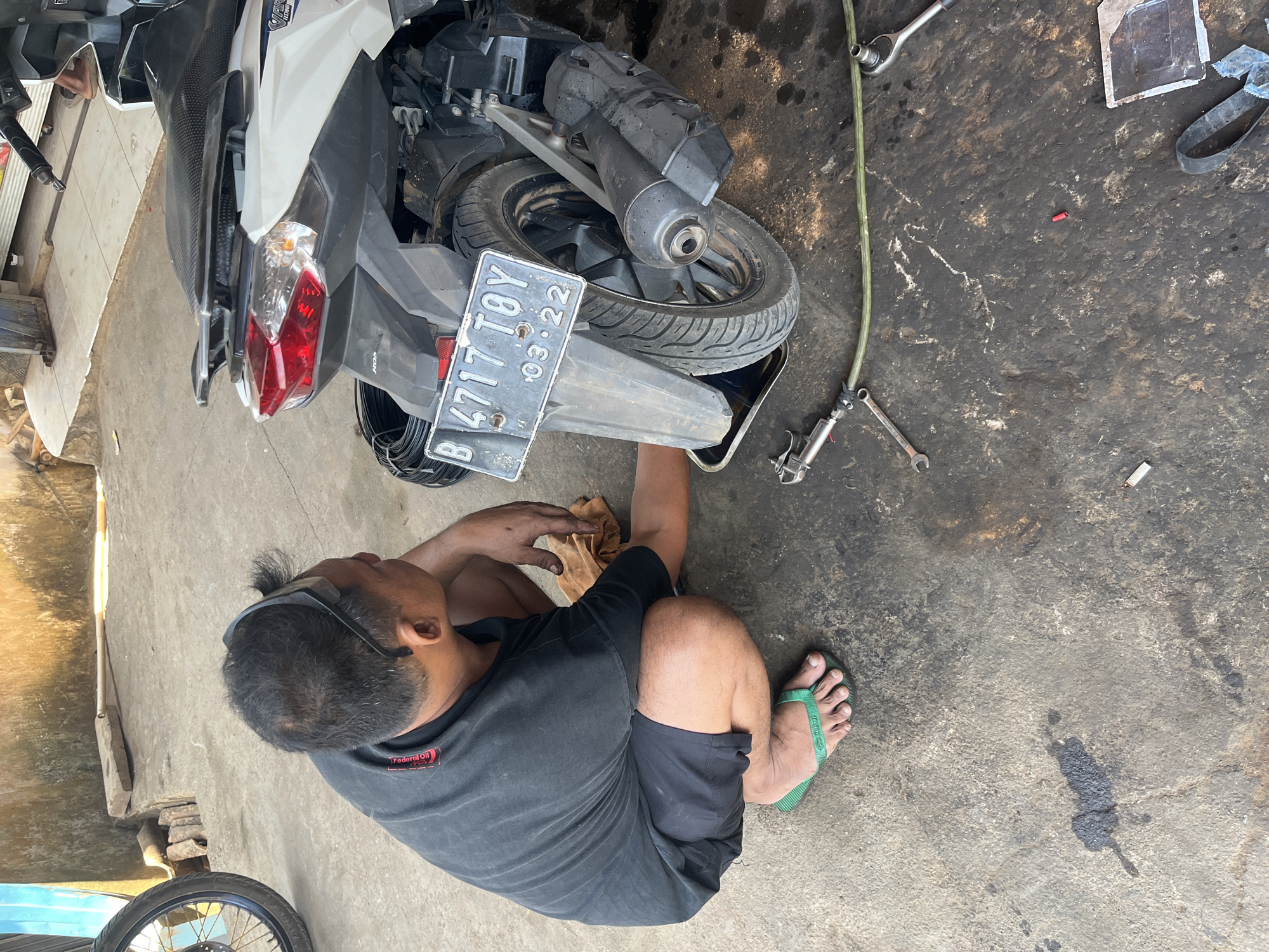 A person is working on a motorcycle in what appears to be a mechanic's shop or a residential area in Karang Sari, Indonesia. The person, wearing a black t-shirt and black shorts, is crouched down, looking at the rear tire of a white and black motorcycle. They have on green flip-flops. The motorcycle has a license plate that reads "B 4717 TOY 03-22". Various tools, including wrenches and an air hose, are scattered on the concrete ground, which is stained with oil and dirt, indicating a work area. In the background, a wall made of rough concrete blocks is visible, along with parts of another motorcycle and building materials. The lighting suggests it is daytime, possibly in the afternoon due to the strong shadows.