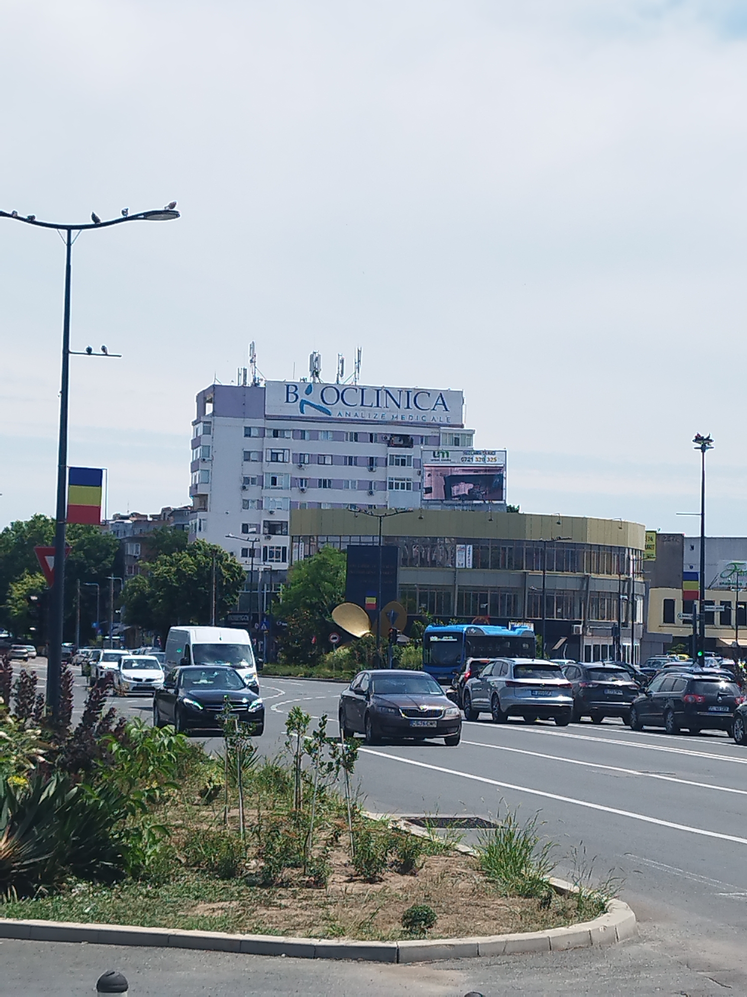 The image depicts a sunny daytime scene on a multi-lane road in Constanța, Romania. In the background, a tall white building is topped with a prominent sign for "BIOCLINICA ANALIZE MEDICALE." Several cars are visible on the road, including a white van, a black sedan with illuminated headlights, and a blue bus. The foreground features a roadside planting area with green vegetation and a concrete curb. A Romanian flag is displayed on a lamppost to the left of the frame. The sky is mostly clear with some faint clouds.