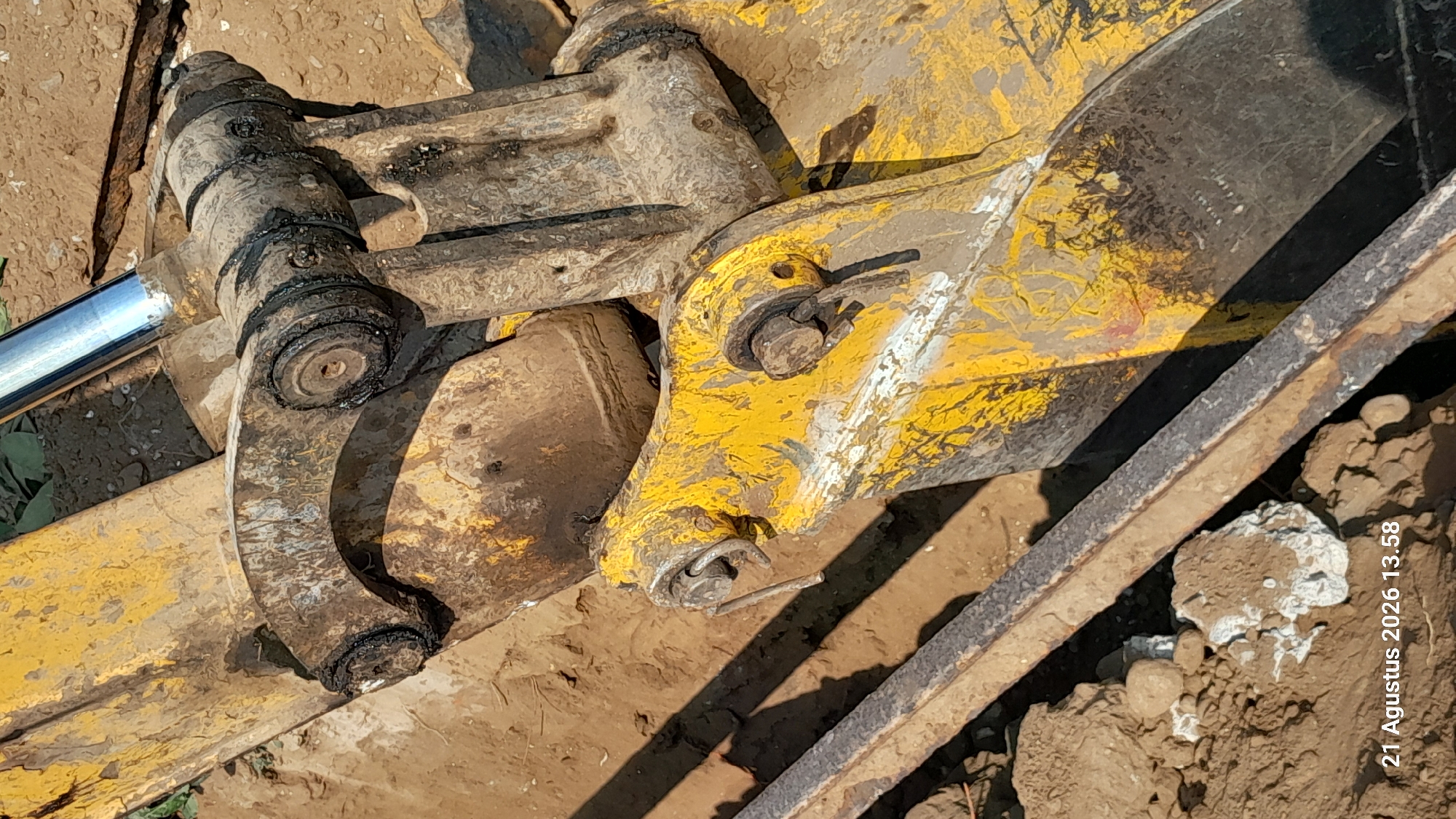 This is a close-up, low-angle, medium shot of the mechanical linkage of a yellow excavator bucket in Takeran, Indonesia. The excavator is positioned on dirt, likely a construction site or excavation area. The focus is on the hydraulic cylinder and the various joints and pins connecting the bucket to the excavator arm. The metal components are worn, dirty, and show signs of grease and rust. The background is blurred, consisting of soil and some green foliage, indicating an outdoor environment. The timestamp "21 Agustus 2026 13.58" is visible in the bottom left corner, suggesting the image was taken on August 21, 2026, at 13:58.