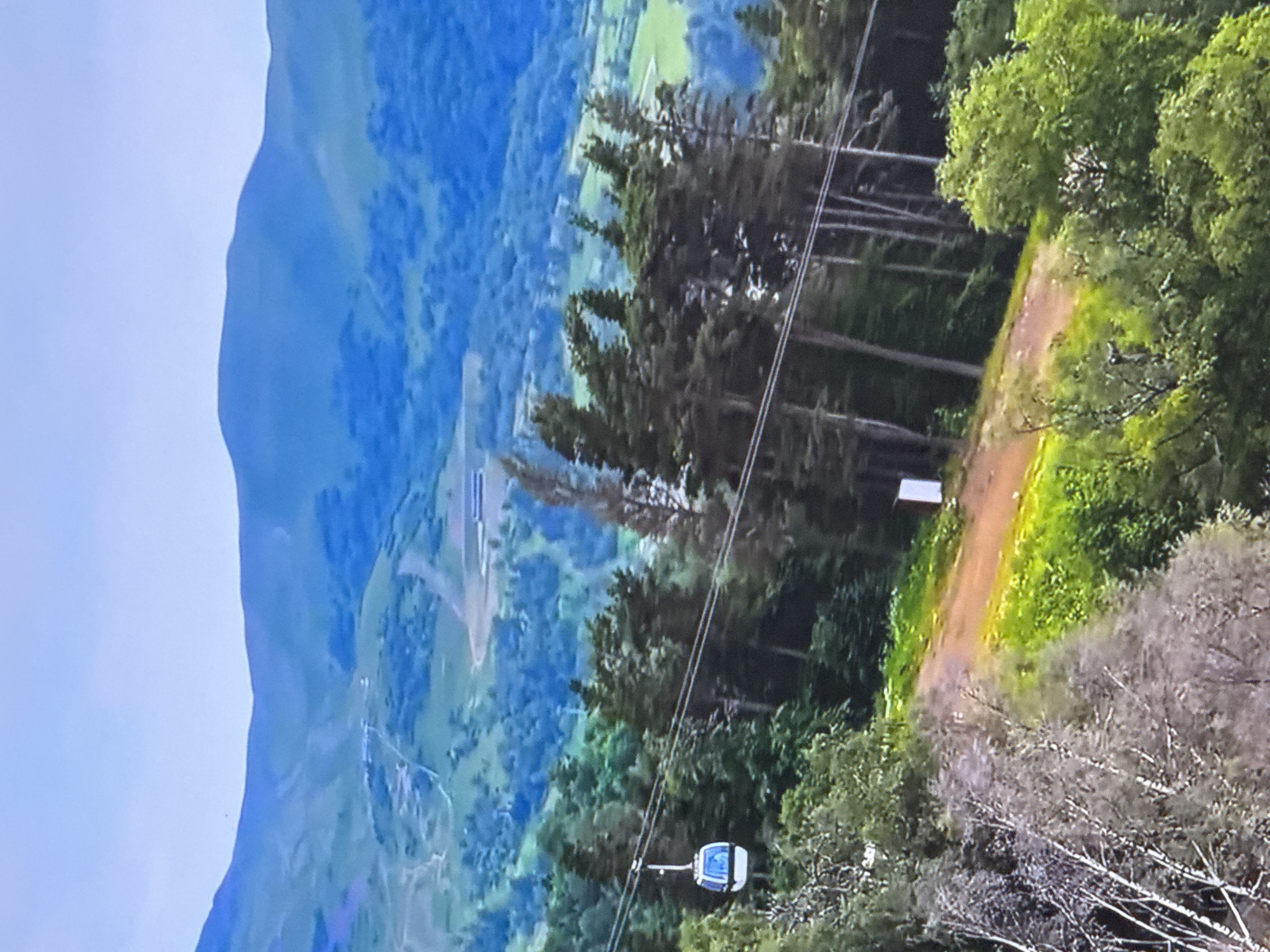 An aerial shot captures a cable car ascending a mountain. The cable car, a white capsule with blue windows, is suspended on a wire against a backdrop of lush green trees and rolling hills. In the distance, a range of blue-hued mountains stretches across the horizon under a clear sky. The foreground features a dirt path winding through verdant foliage.