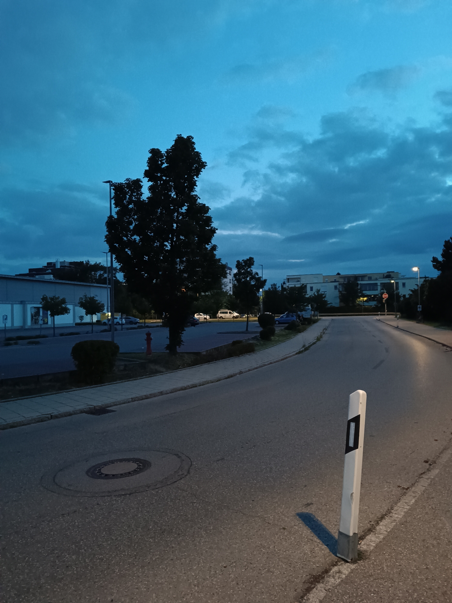 The image depicts an exterior street scene in Germering, Germany, during twilight. A paved road curves to the right, bordered by a sidewalk on the left and a grassy median with a white traffic marker on the right. A manhole cover is visible on the road surface. In the background, buildings and trees are silhouetted against a deep blue sky with scattered clouds. Several vehicles are parked or driving on the road. A red fire hydrant stands near a tree on the left side of the image. The lighting suggests dusk or early evening.
