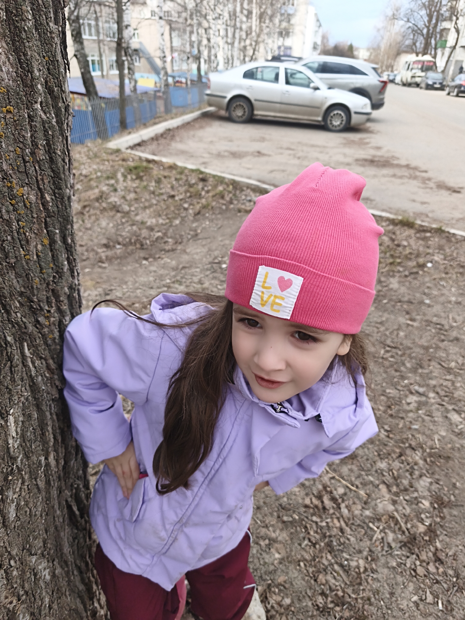 A young girl is depicted outdoors, playfully leaning against a large tree trunk and looking up towards the viewer with a slight smile. She is wearing a vibrant pink knit beanie hat with a white rectangular label on the front, featuring the word "L❤️VE" in yellow lettering. Her long, dark hair peeks out from under her hat, and she is dressed in a light purple, possibly waterproof, jacket, with maroon or dark red pants visible below. Her hands appear to be on her hips or tucked into her pockets, adding to her casual pose.
The tree trunk on the left side of the frame features rough, dark bark with patches of yellow-green moss or lichen. The ground around the girl is covered with dry, fallen leaves and small twigs, suggesting a setting in late autumn, winter, or early spring.
In the blurred background, there's an urban or suburban street with several parked cars, including a silver sedan and a grey SUV. Residential buildings with light-colored facades are visible behind the vehicles, partially obscured by bare-branched trees. A blue picket fence also runs along a section of the background. The sky is overcast, indicating a daytime scene with soft, diffused light.