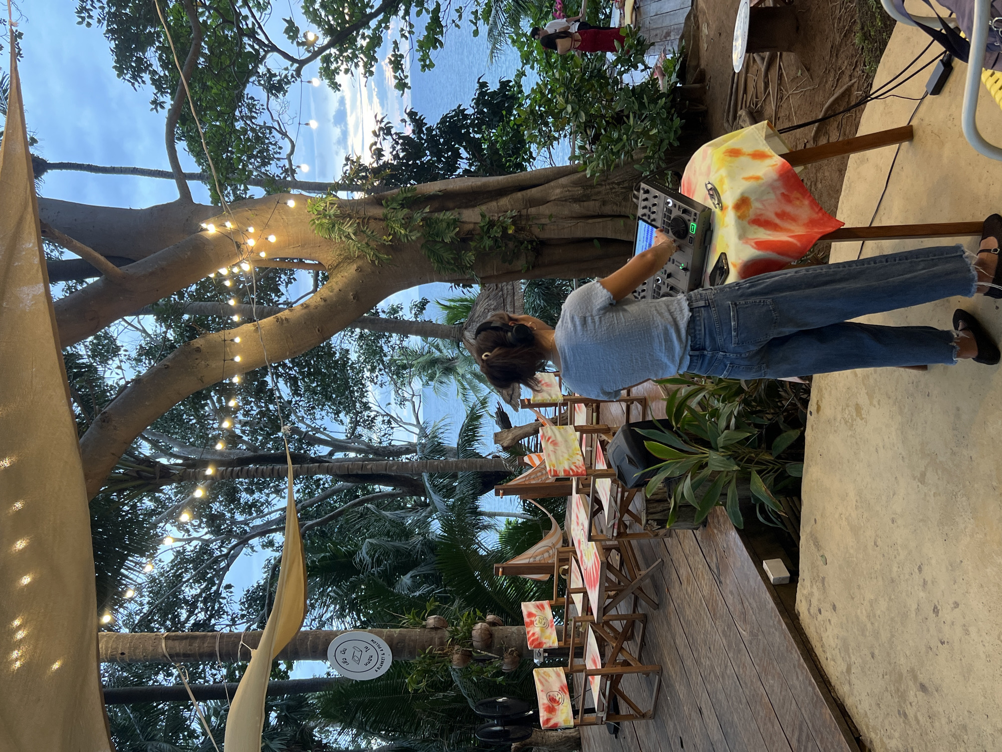 The image captures an outdoor scene at what appears to be a beachside venue in Karon, Thailand. A person, wearing headphones and a light blue shirt with jeans, is standing and operating a DJ mixer. The DJ mixer is placed on a table adorned with a tie-dye fabric in shades of red, yellow, and white. In the background, lush green trees and palm trees dominate the view, with a glimpse of the ocean and a cloudy sky suggesting late afternoon or early evening. String lights are visible, adding to the ambiance of the setting. Several wooden chairs with similar tie-dye fabric are arranged on a wooden deck, indicating a seating area for guests. A large, imposing tree trunk is a prominent feature in the scene. A round sign with Thai text is attached to a palm tree trunk, though the text is not clearly legible. The overall atmosphere suggests a relaxed, possibly festive, setting, perhaps a bar or restaurant by the sea.