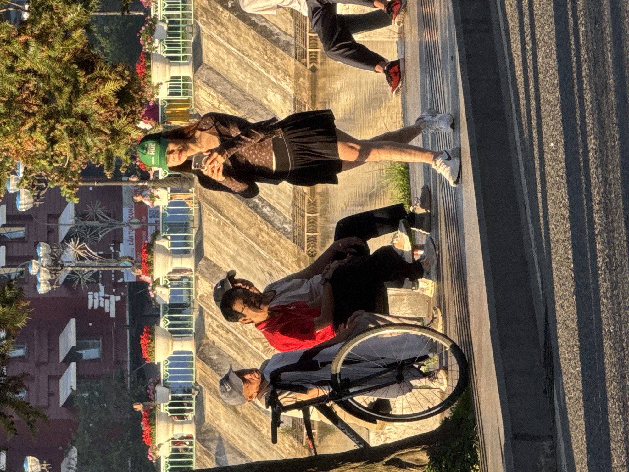 The image captures a scene in Dalat, Vietnam, likely near Xuan Huong Lake, given the presence of a paved walkway, a body of water, and decorative planters with flowers. In the foreground, a black bicycle is parked next to a tree. To the left, three men are seated on a bench. One man wears a grey baseball cap and a light-colored shirt, another wears a red polo shirt and black pants, and the third, partially obscured, wears a dark cap and a white shirt. In the center and right of the frame, a young woman stands, holding a smartphone. She wears a dark, sheer patterned long-sleeved top over a patterned undergarment, a black pleated skirt, and white athletic shoes. She is also wearing a green baseball cap with the word "HUE" printed on it. In the background, a low wall with decorative white planters filled with red flowers separates the walkway from a building with a dark red facade. The setting sun casts long shadows across the paved area.