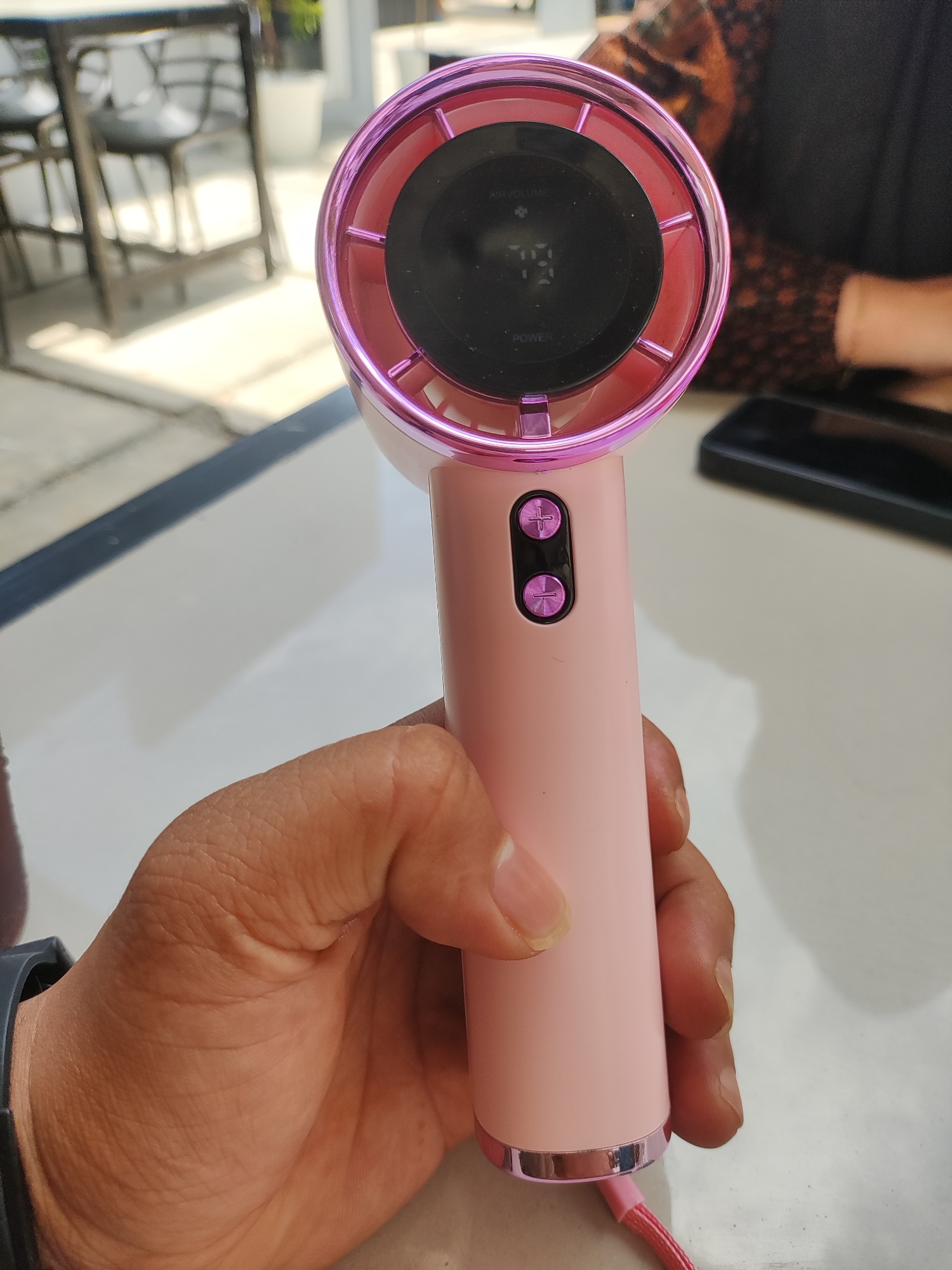 A hand holds a pink portable fan with a circular head, featuring a digital display showing the number "79" and buttons marked with "+" and "-" symbols. The fan has a metallic pink rim and a matching pink handle with a silver accent at the base. The fan is on a table. Part of a person's arm and clothing is visible in the background, along with a black electronic device and a table. In the blurred background, chairs, a table, and greenery are visible. The location is inferred to be in Lhokseumawe, Indonesia.
