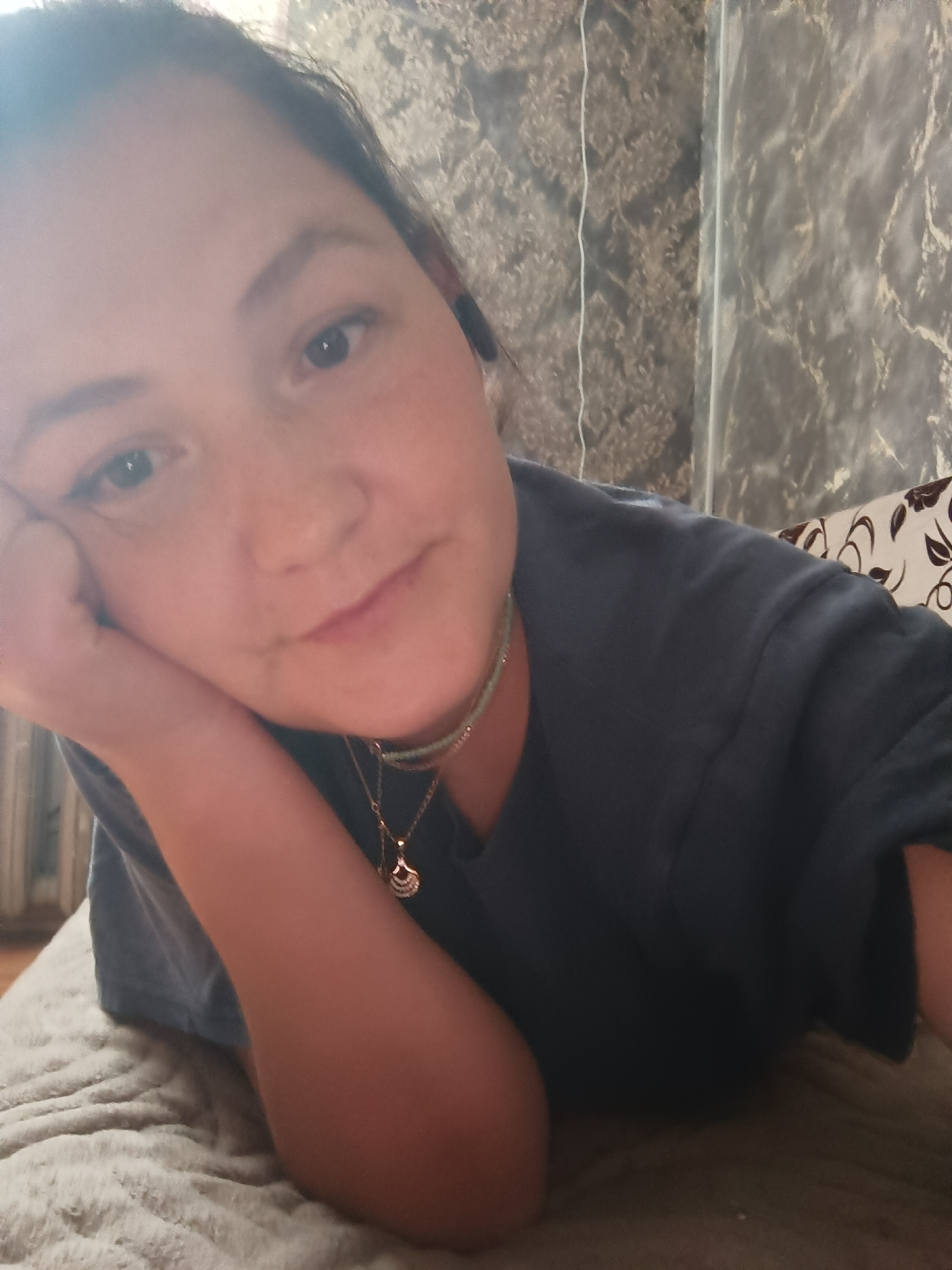 A young woman with dark hair is lying on her side, propped up on her elbow, and looking directly at the camera. She has her hand resting on her cheek and is wearing a dark blue t-shirt and a necklace. The background appears to be a wall with patterned wallpaper and part of a bed with a textured blanket. The lighting is soft and appears to be natural or indoor lighting. The image was taken in an indoor setting, likely a bedroom, in the city of Karsha, Russia.