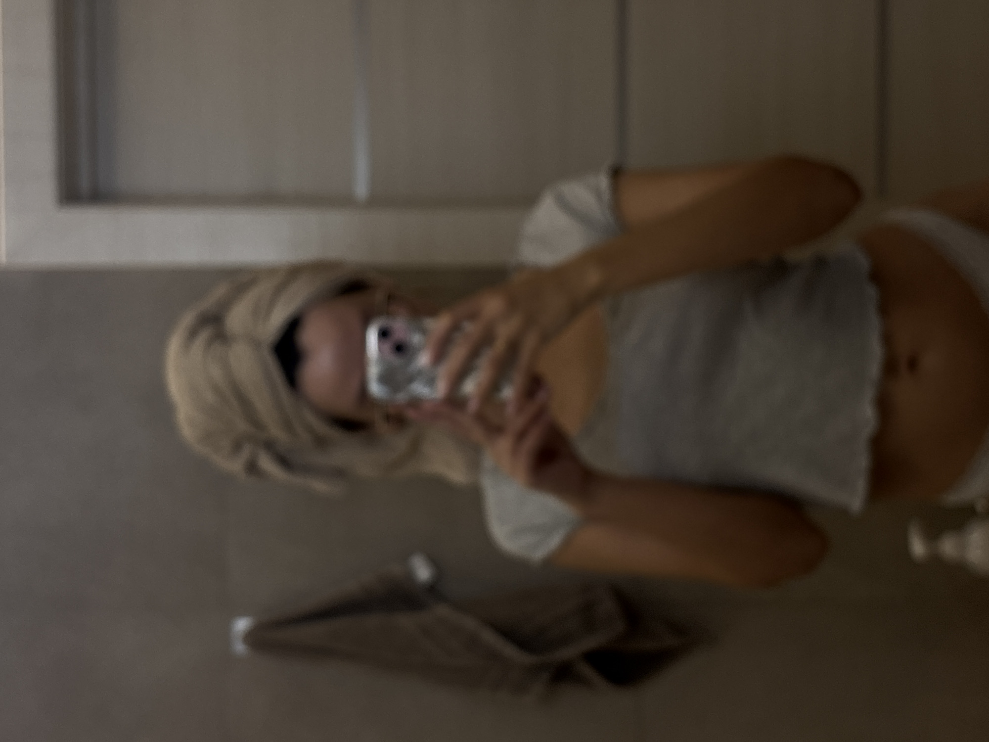 A blurry, rotated image captures a person taking a mirror selfie indoors. The subject, likely female, has blonde hair that appears damp or wrapped, possibly post-shower. She is wearing a light-colored, short-sleeved crop top that exposes her midriff, and light-colored bottoms. She holds a light-colored smartphone, possibly an iPhone in a clear case, to take the photograph, with the phone obscuring her face.
The setting appears to be a bathroom, characterized by a mirror reflection and a dark-colored towel hanging on a hook or rack on the wall below the person's torso. To the right of her lower body, a small, light-colored bottle, possibly for soap or lotion, rests on a surface. The background features light grey or beige walls with what looks like cabinet doors or wall panels above. The lighting is dim. Due to the blurriness and angle, no text is visible on any objects.