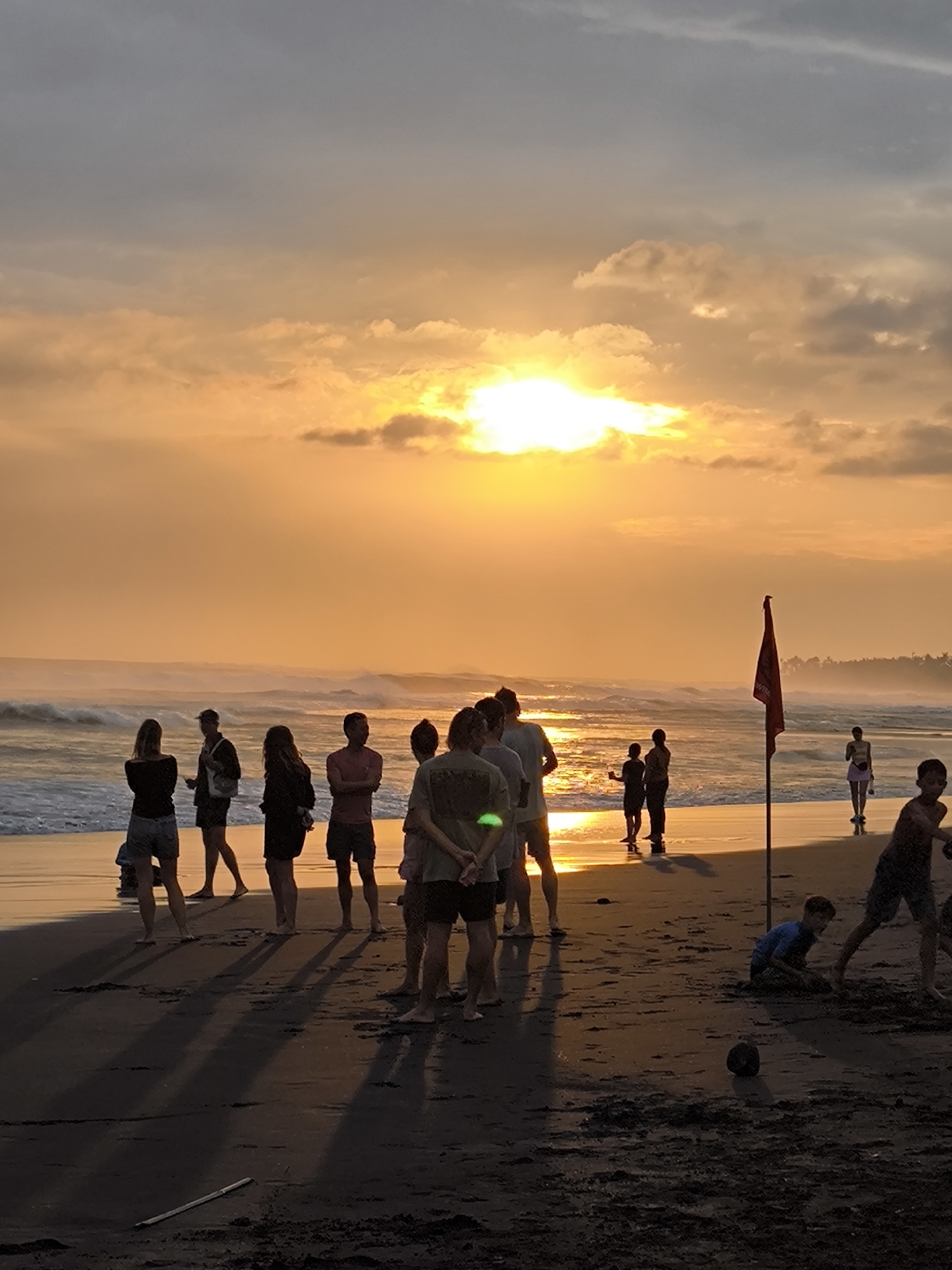 A group of people are gathered on a beach at sunset in North Kuta, Indonesia. The sky is a vibrant orange and yellow, with some grey clouds. The sun is a bright orb near the horizon, casting long shadows across the dark sand. Waves are rolling in from the ocean, creating a foamy edge on the shore.
Several individuals are standing and facing the ocean, some are talking to each other. In the foreground, a young boy in a blue shirt is crouching on the sand, appearing to be playing with something. Further back, a red flag on a pole stands out against the sunset. The overall mood of the scene is peaceful and relaxed, with people enjoying the natural beauty of the beach at dusk. The clothing worn by the people suggests a warm climate, consistent with the tropical location.