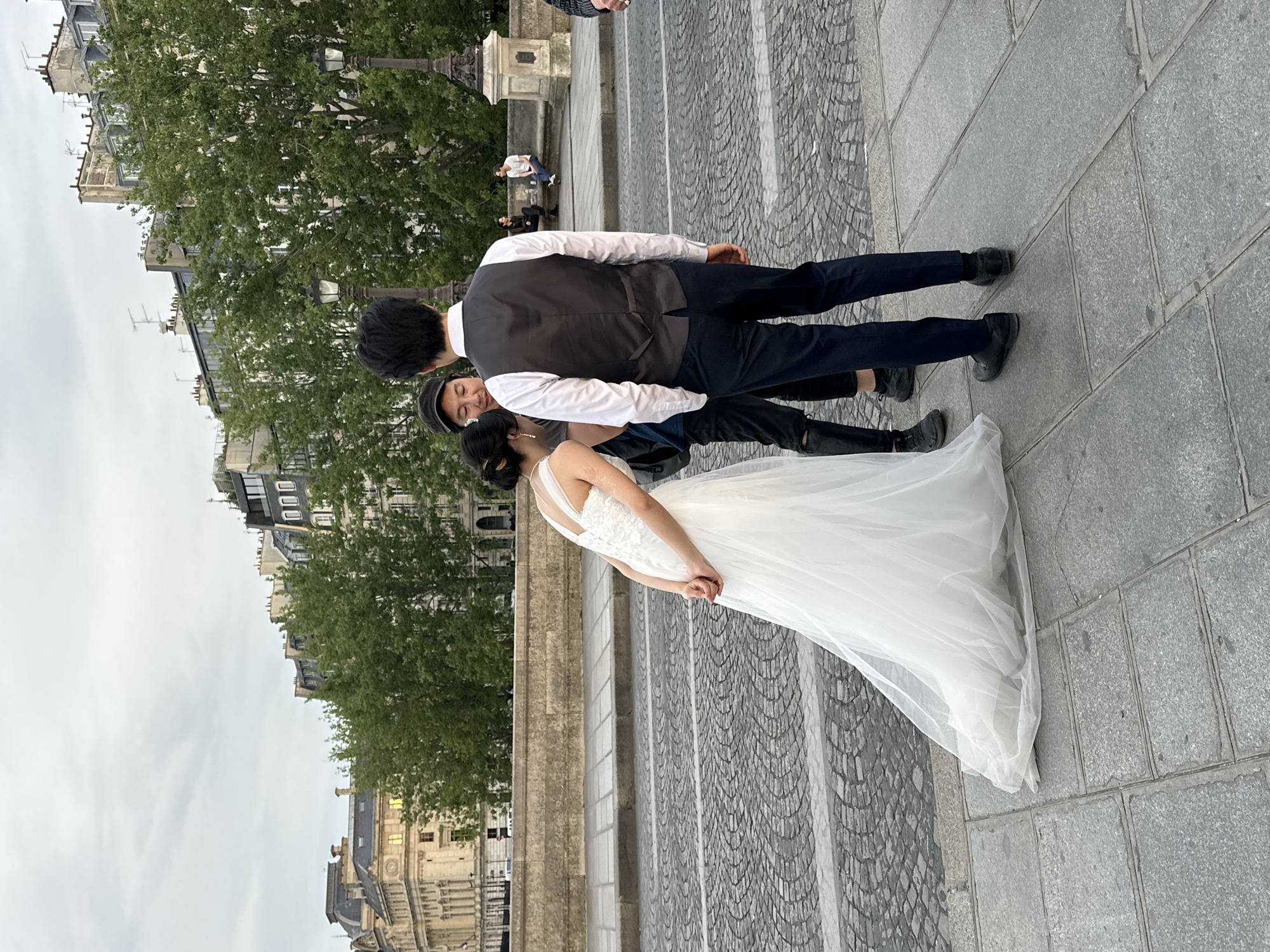 A couple, possibly newly married, is posing for a photo in Paris, France. The bride is wearing a white wedding gown and the groom is dressed in black trousers, shoes, and a black vest over a white shirt. A third individual wearing a hat stands next to the bride and groom. The photograph is taken on a bridge with visible cobblestone pavement and stone steps. Buildings and trees can be seen in the background, typical of the Parisian landscape. Other people are sitting or standing further in the background. The sky appears overcast.