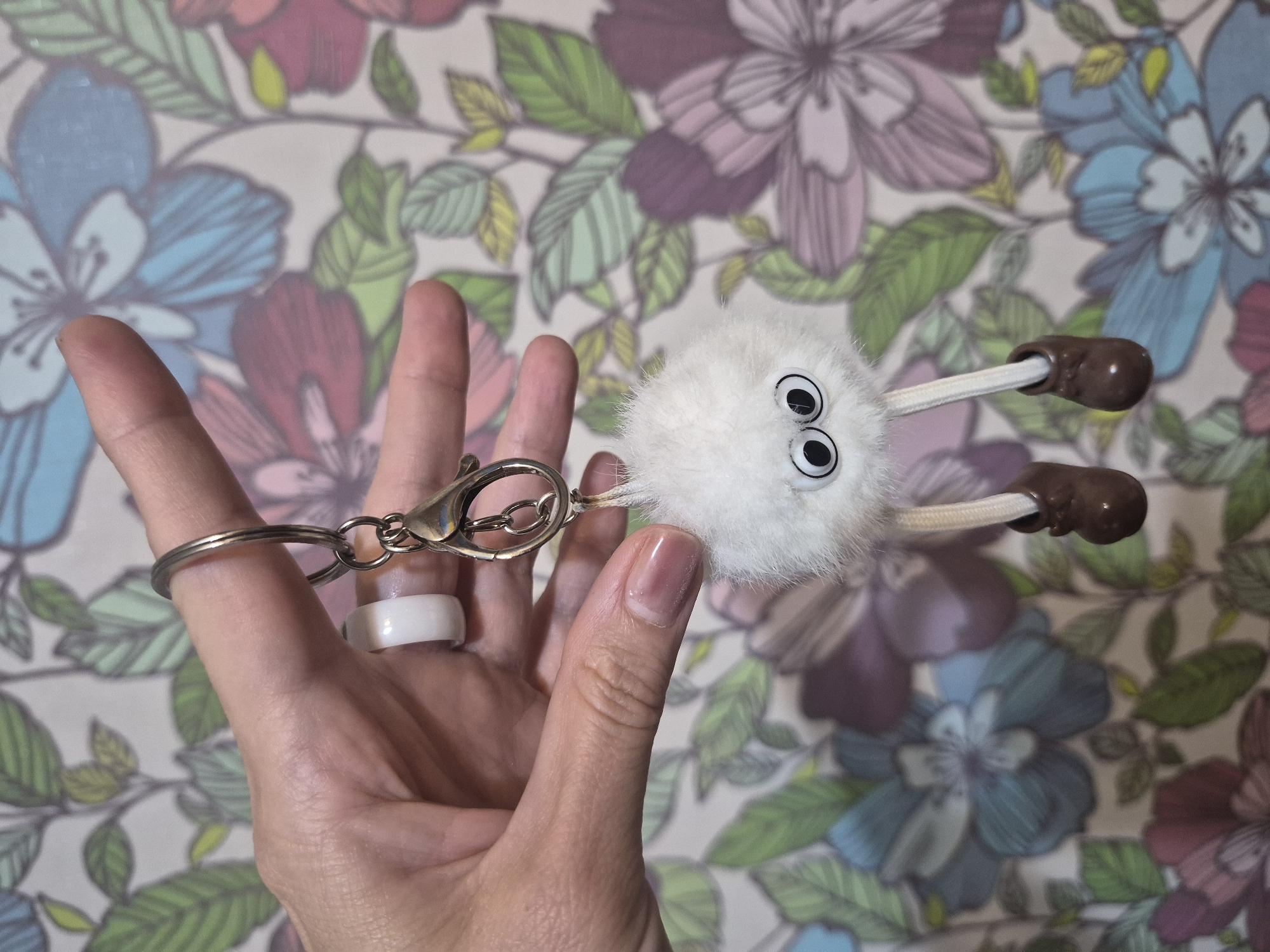A hand with fingernail polish is holding a white fuzzy keychain with googly eyes and brown shoes. The background is a wallpaper with a floral pattern. The image does not contain information about the location in the city of Chaikovsky, Russia.